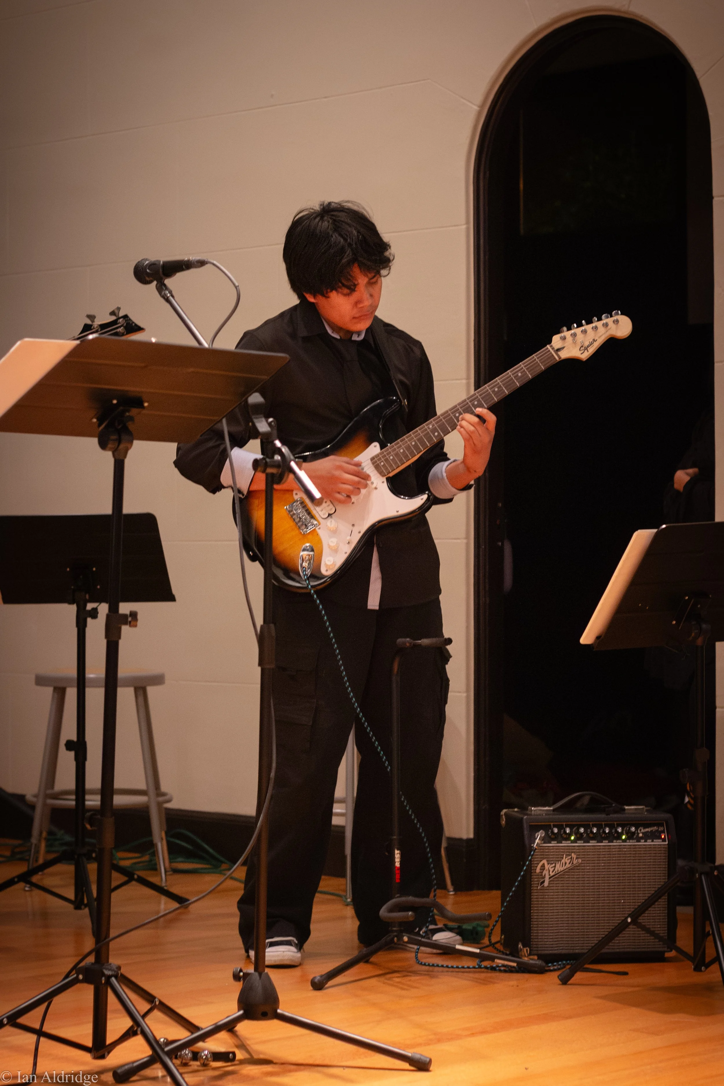 A young man playing an electric guitar on stage, with music stands and a Fender amplifier nearby.
