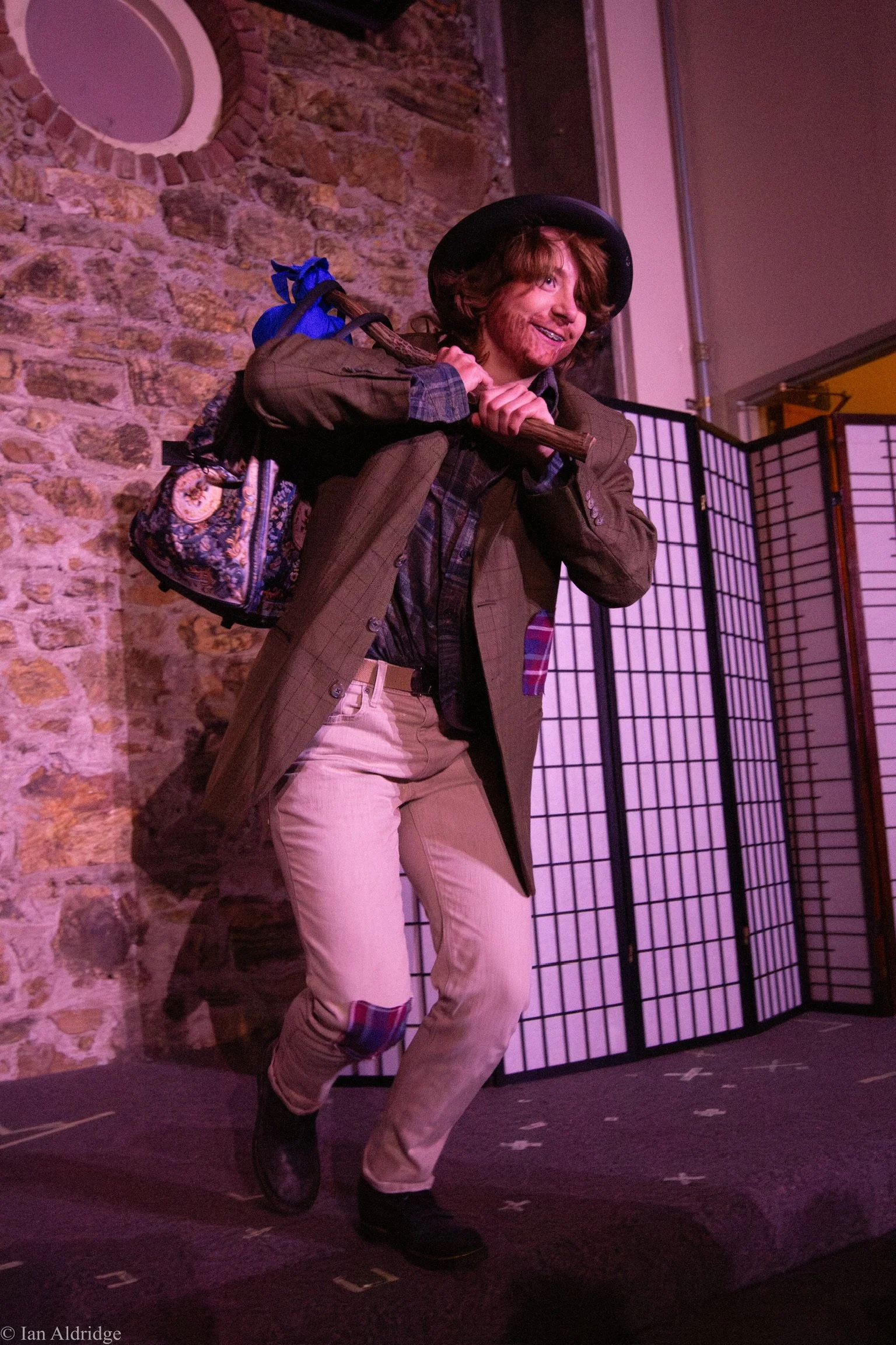 A man dressed in vintage clothing, including a plaid shirt, a blazer, beige pants, and a black hat, holding an axe over his shoulder, smiling and standing in front of a brick wall and a folding room divider.