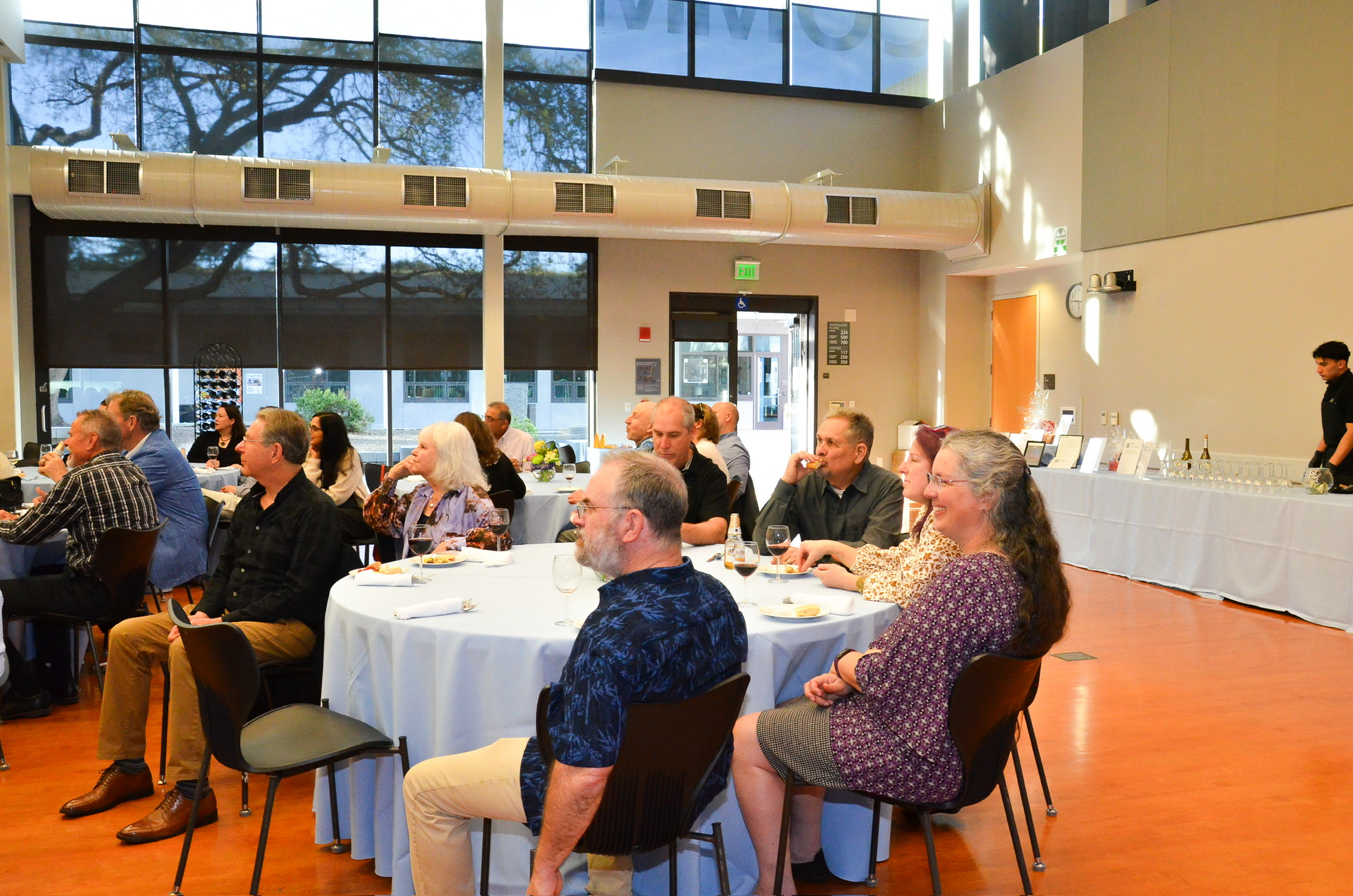 A diverse group of people seated at round tables during a formal event in a bright, spacious room with large windows. Some attendees are eating and drinking, enjoying the occasion.
