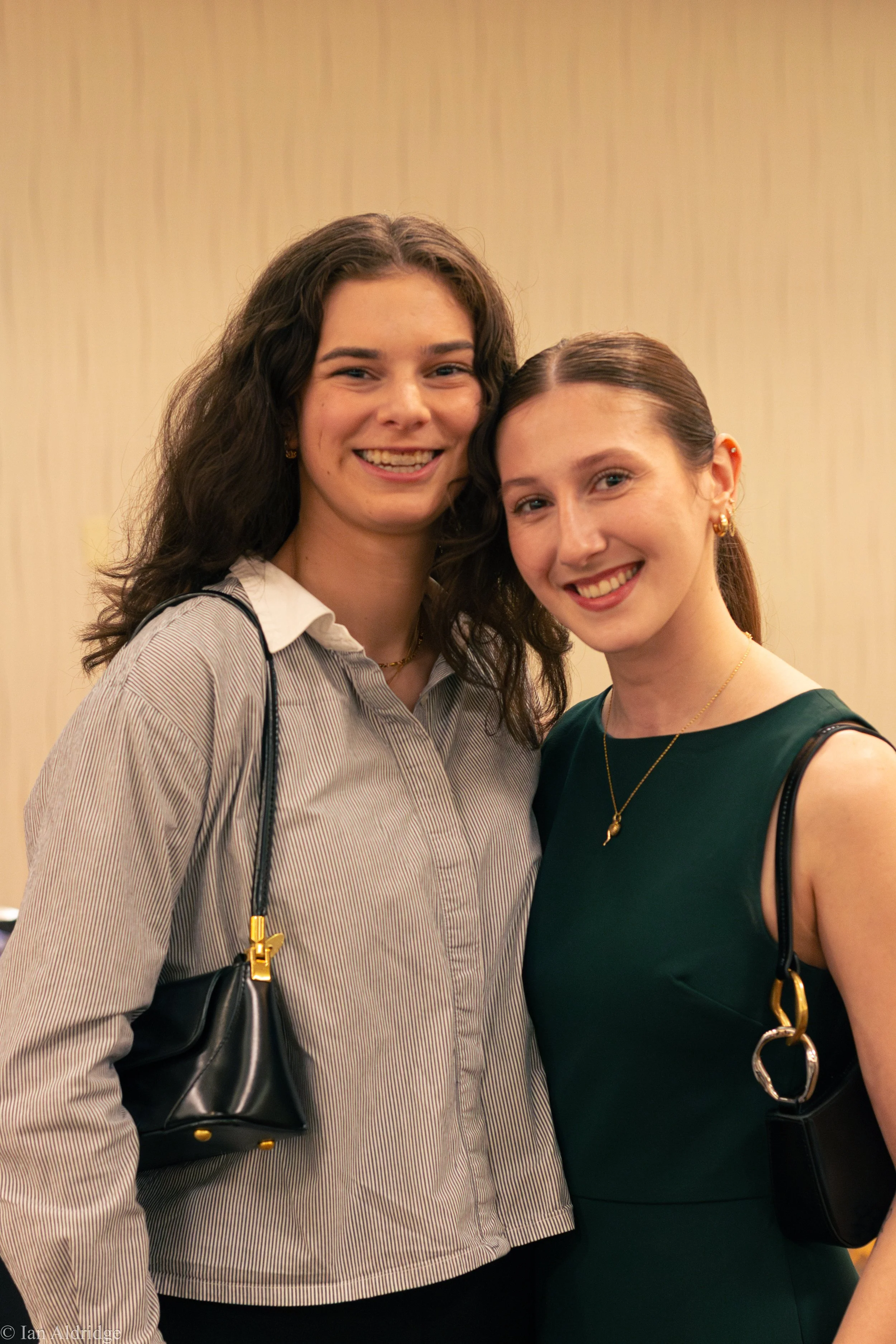 Two women smiling and standing close together at an indoor event, with a plain beige wall background. One woman has long dark wavy hair, wearing a striped button-up shirt, and carrying a black purse. The other woman has straight brown hair tied back,