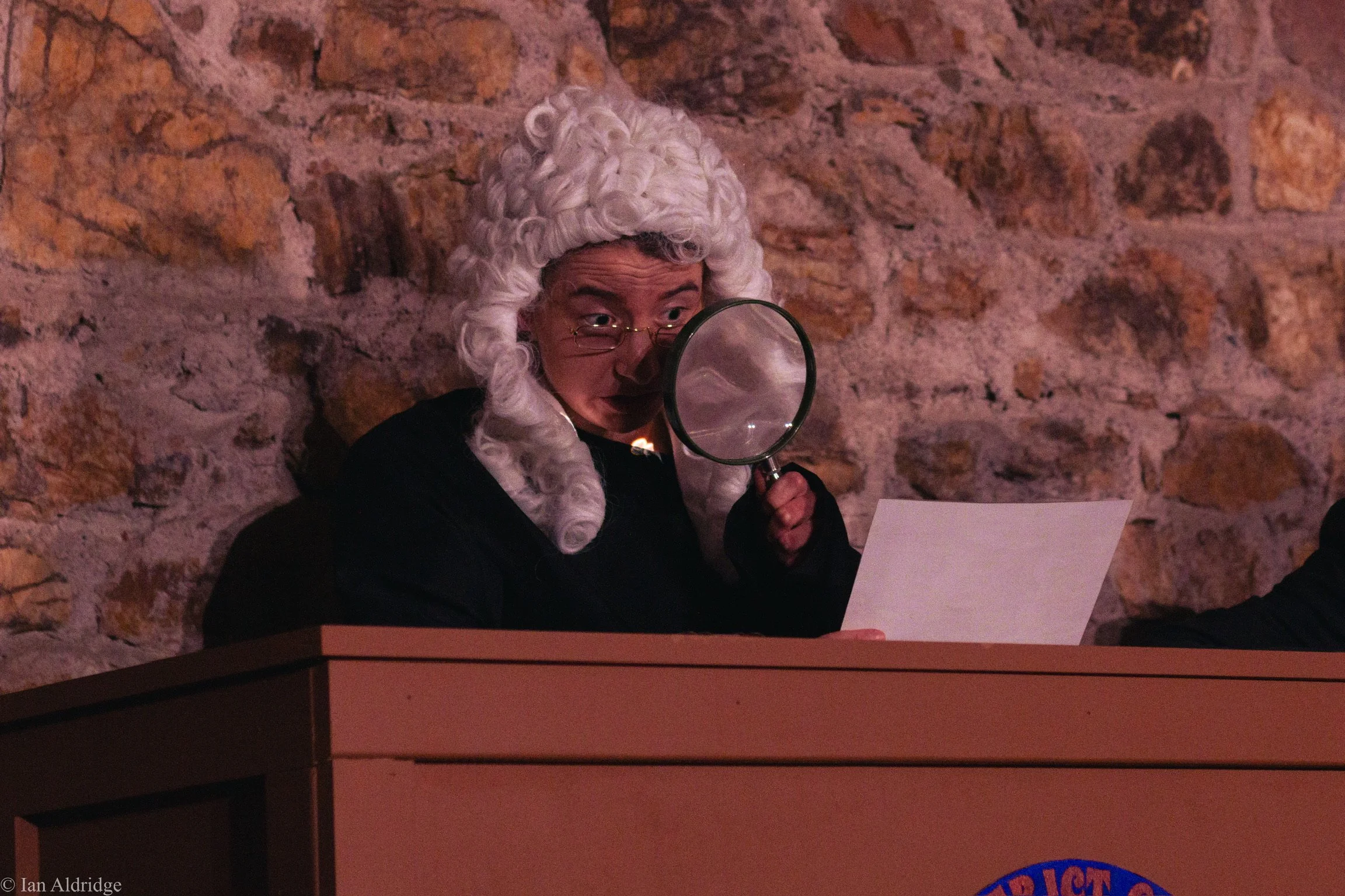Person dressed as a judge with a white curly wig and glasses, holding a magnifying glass near their face, sitting at a table against a stone wall background.