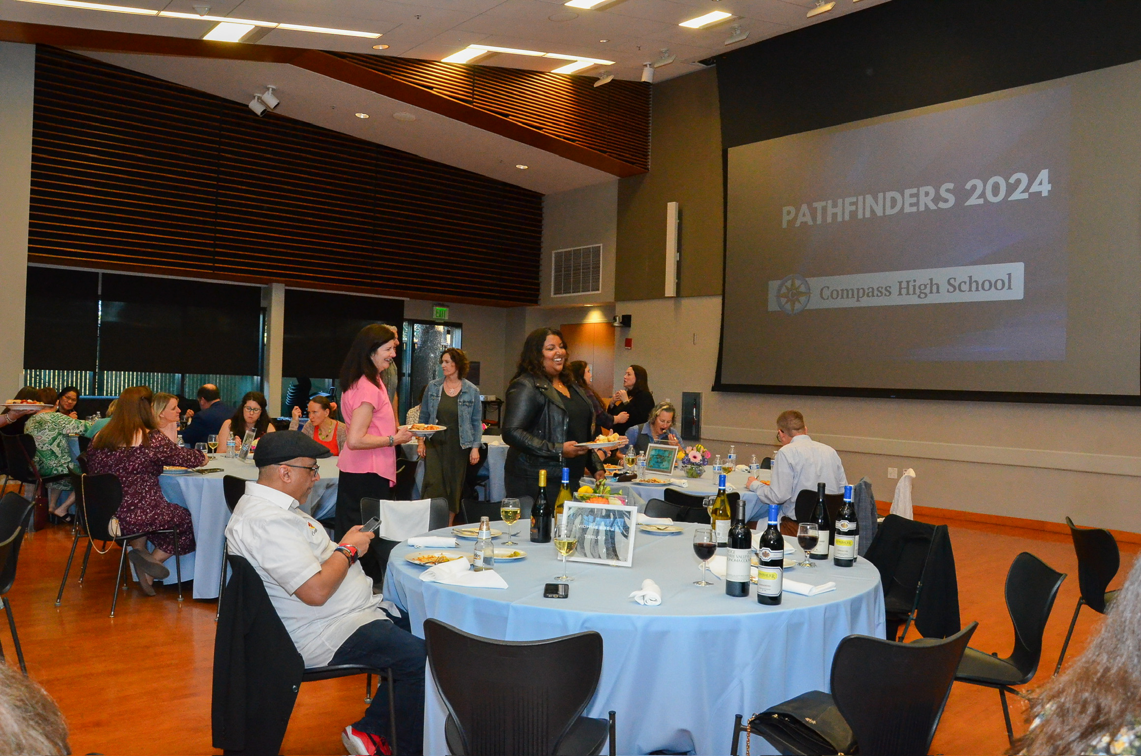 People attending an event at Compass High School with a presentation slide that reads "PATHFINDERS 2024" on a large screen. The room has round tables set with wine bottles, glasses, and floral centerpieces, and attendees are seated or standing, engag