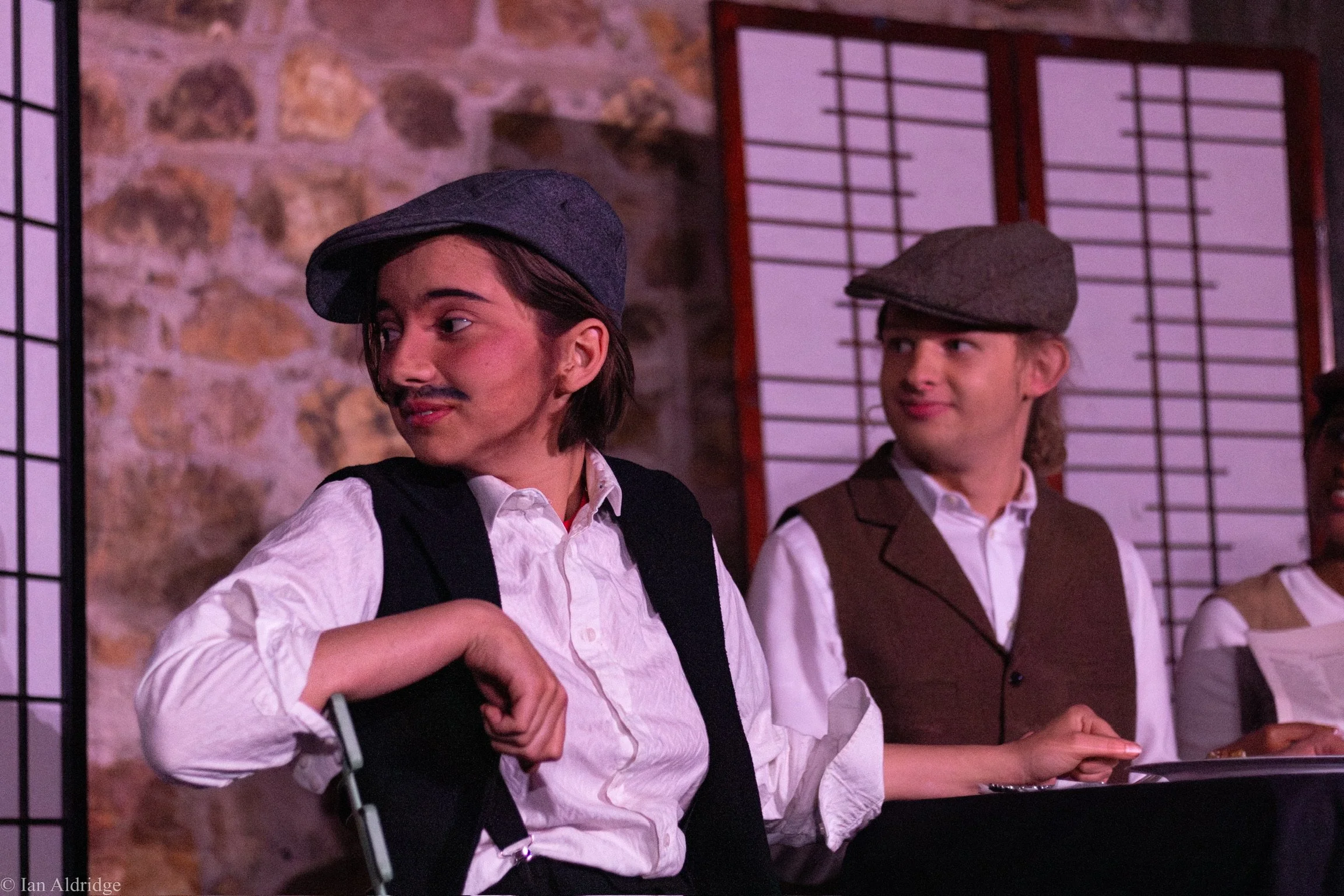 Two young men dressed in vintage attire, one wearing a gray cap and suspenders, and the other wearing a gray beret and vest, standing in front of a stone wall and shoji screen, looking at each other.