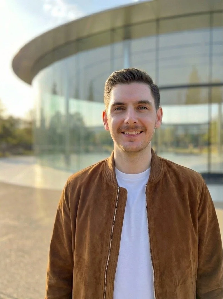 Smiling person wearing a black sweater and standing outdoors with blurred trees in the background.