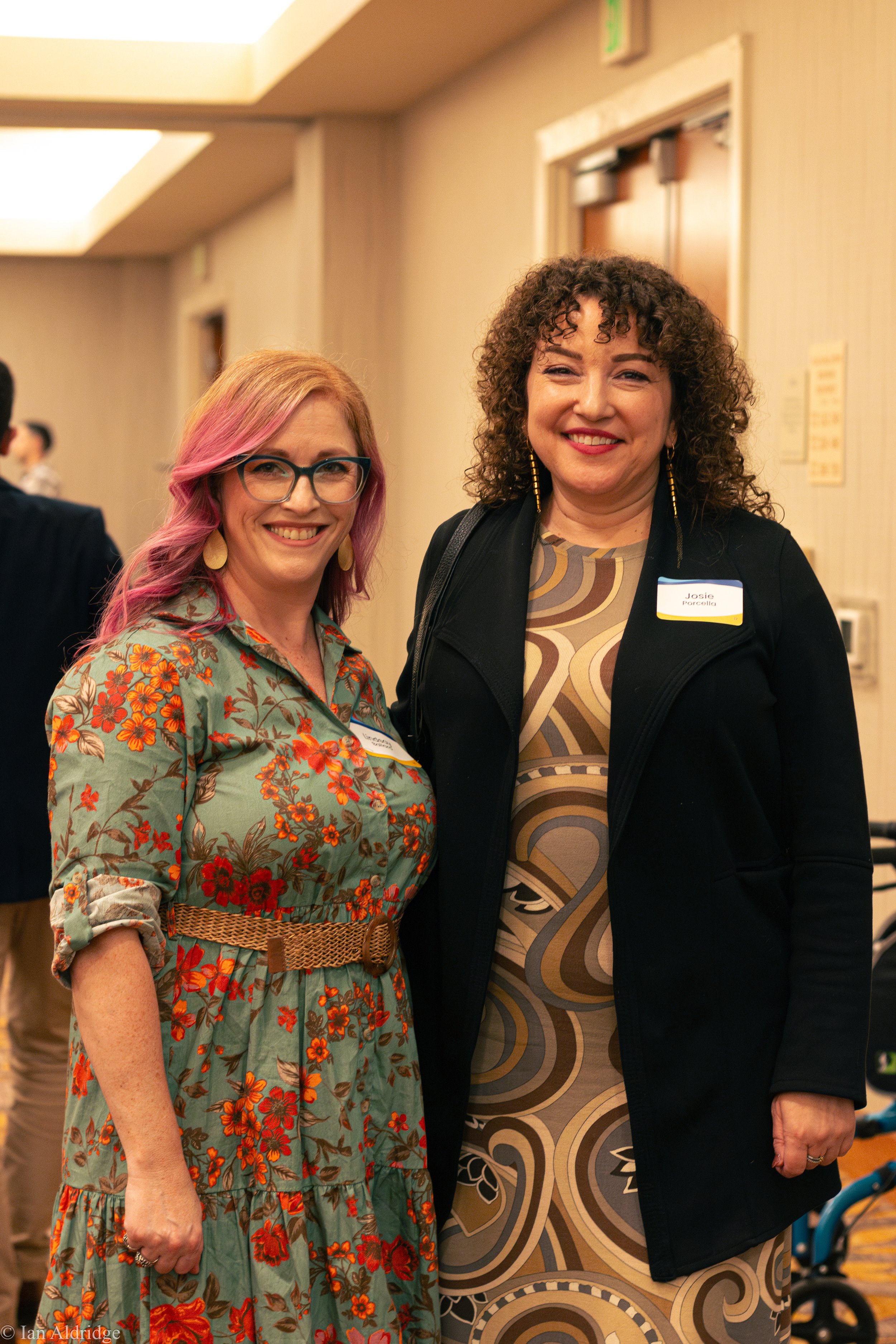 Two women smiling at an indoor event. The woman on the left has pink hair, glasses, and is wearing a floral dress. The woman on the right has curly dark hair, earrings, and is wearing a patterned dress with a black blazer. Both have name tags.