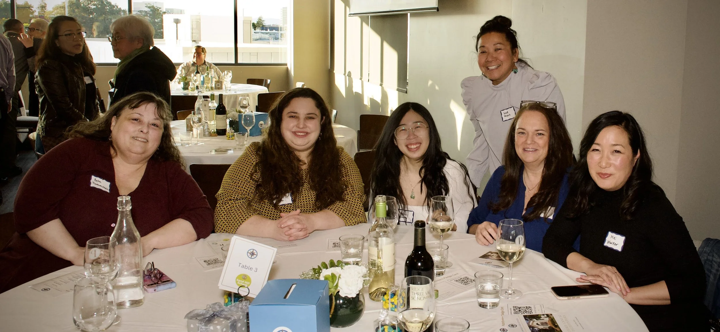 Group of five women sitting at a round table in a well-lit banquet room, smiling at the camera. The table is set with wine glasses, bottles, and floral centerpieces, indicating a social or formal event.