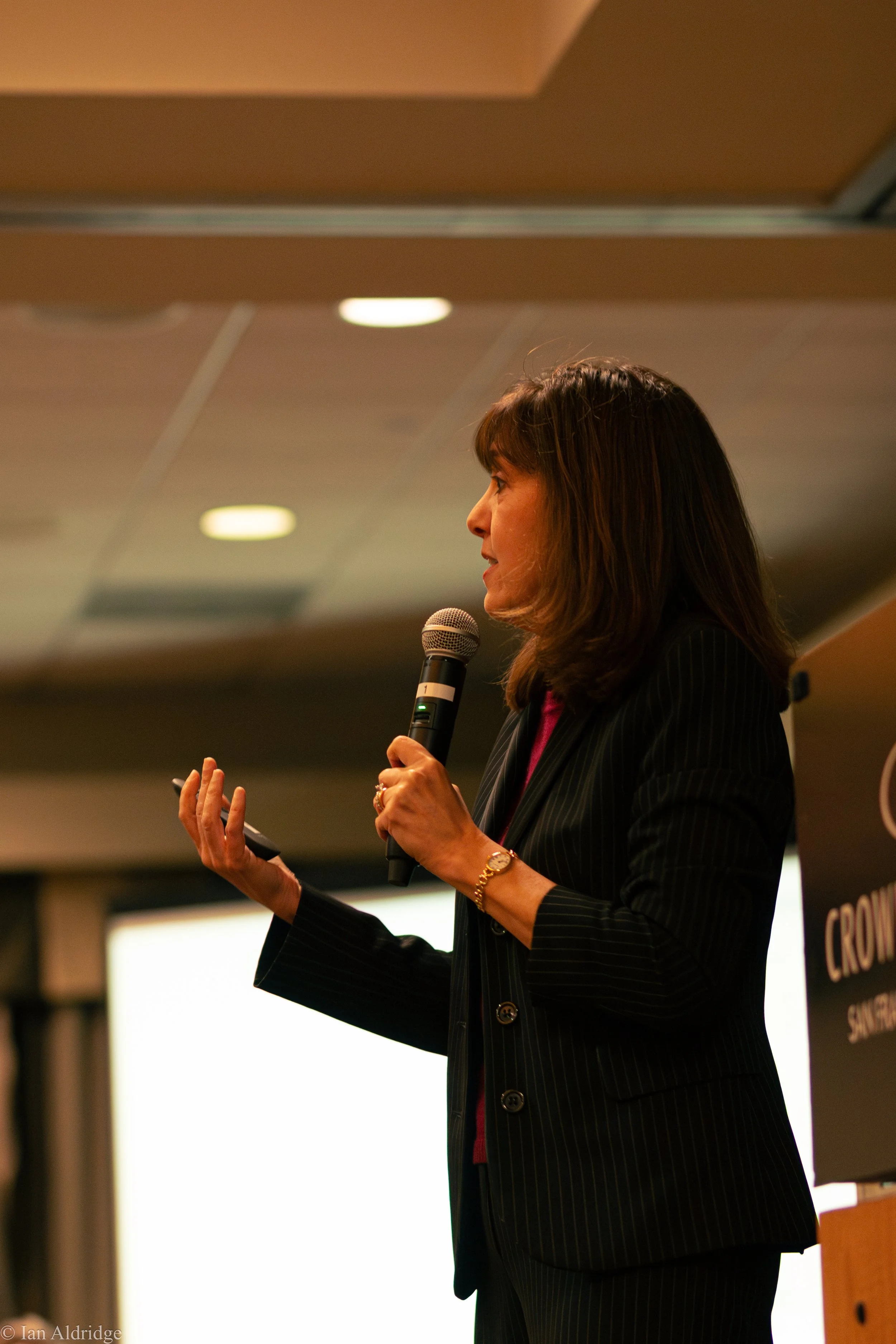 Side view of a woman in a dark pinstripe suit holding a microphone and speaking at a conference or event.