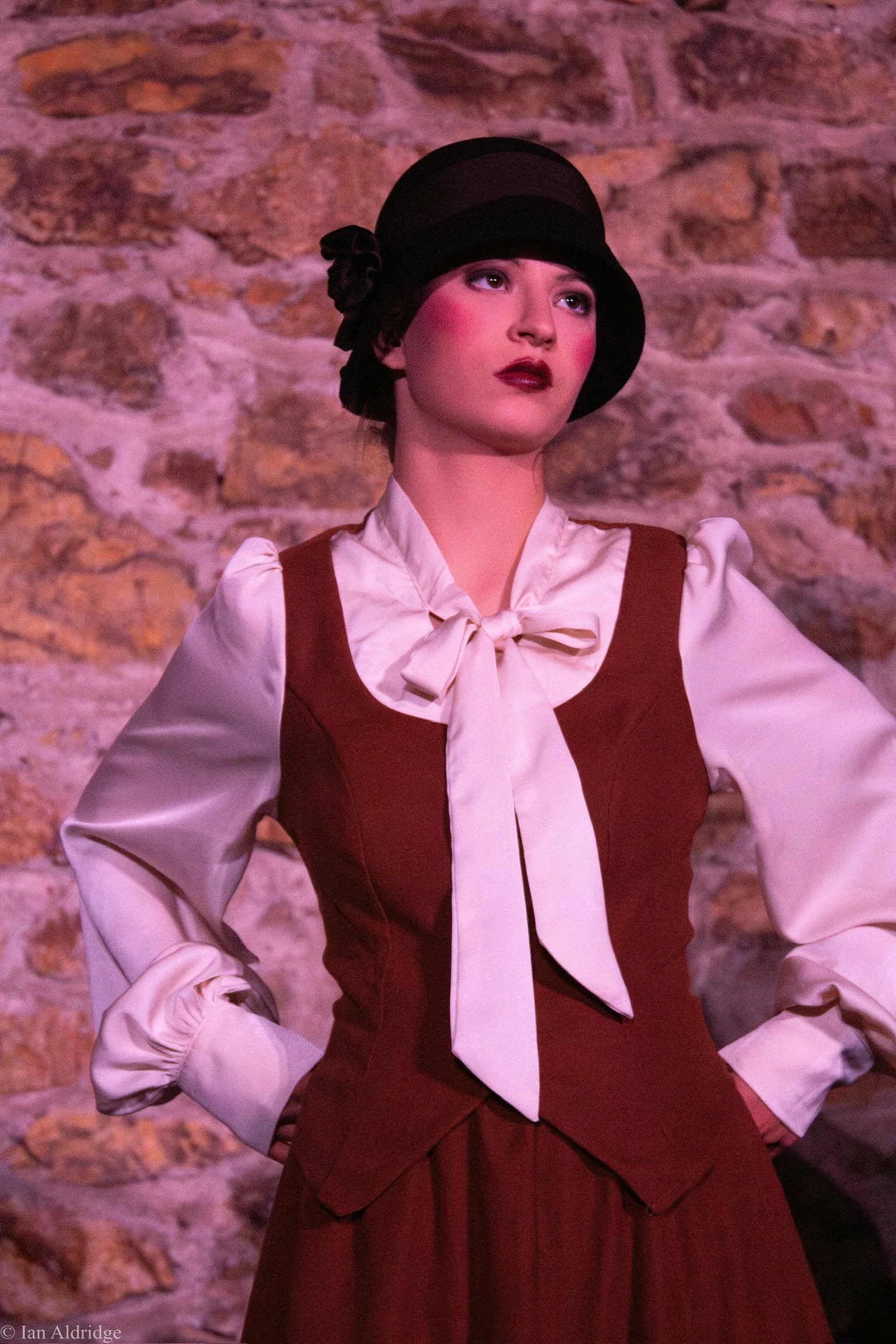 A woman dressed in vintage style clothing with a white blouse, red vest, and a matching long red skirt, wearing a black hat with a flower, standing against a stone wall.