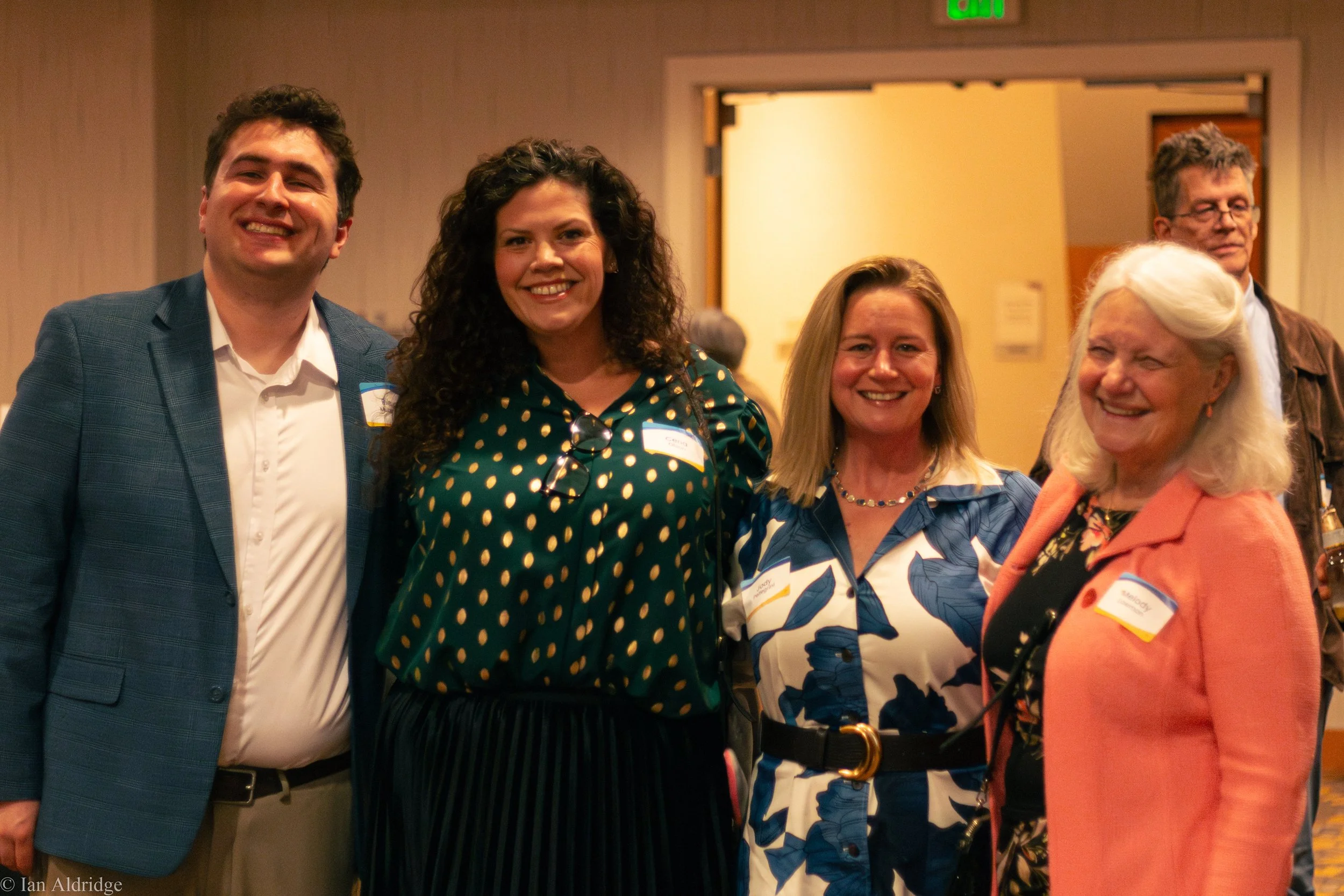 Group of five smiling people attending an event, standing indoors, with name tags on their clothing.
