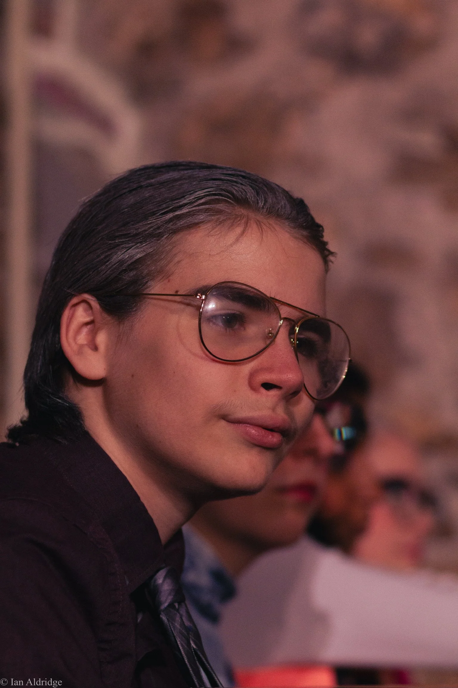 A person with short, dark, slicked-back hair and round glasses looking sideways in a setting with blurred people in the background.