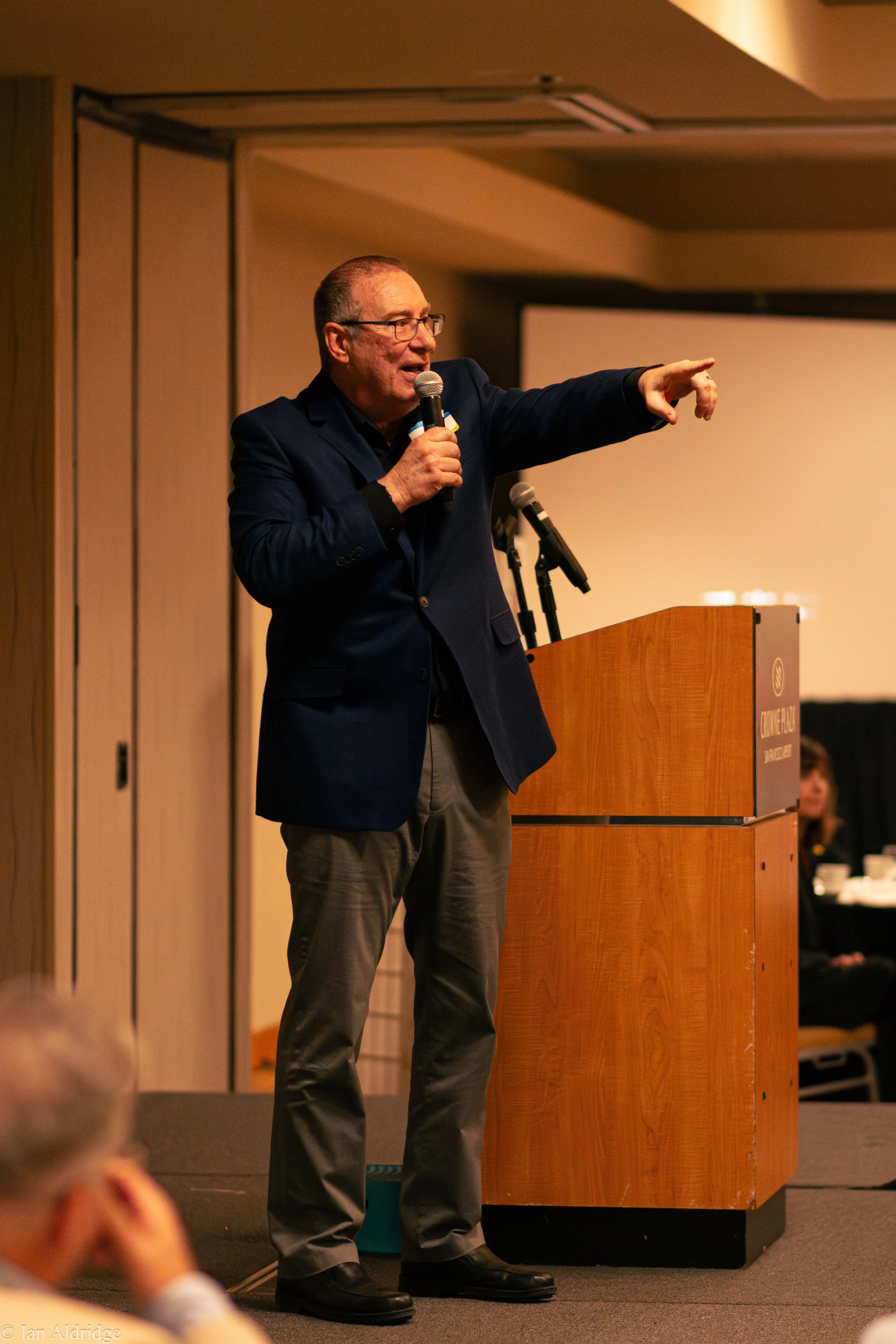 A man in glasses and a dark blazer giving a speech at a podium, gesturing with his right arm extended and pointing. The background shows part of an indoor conference room.