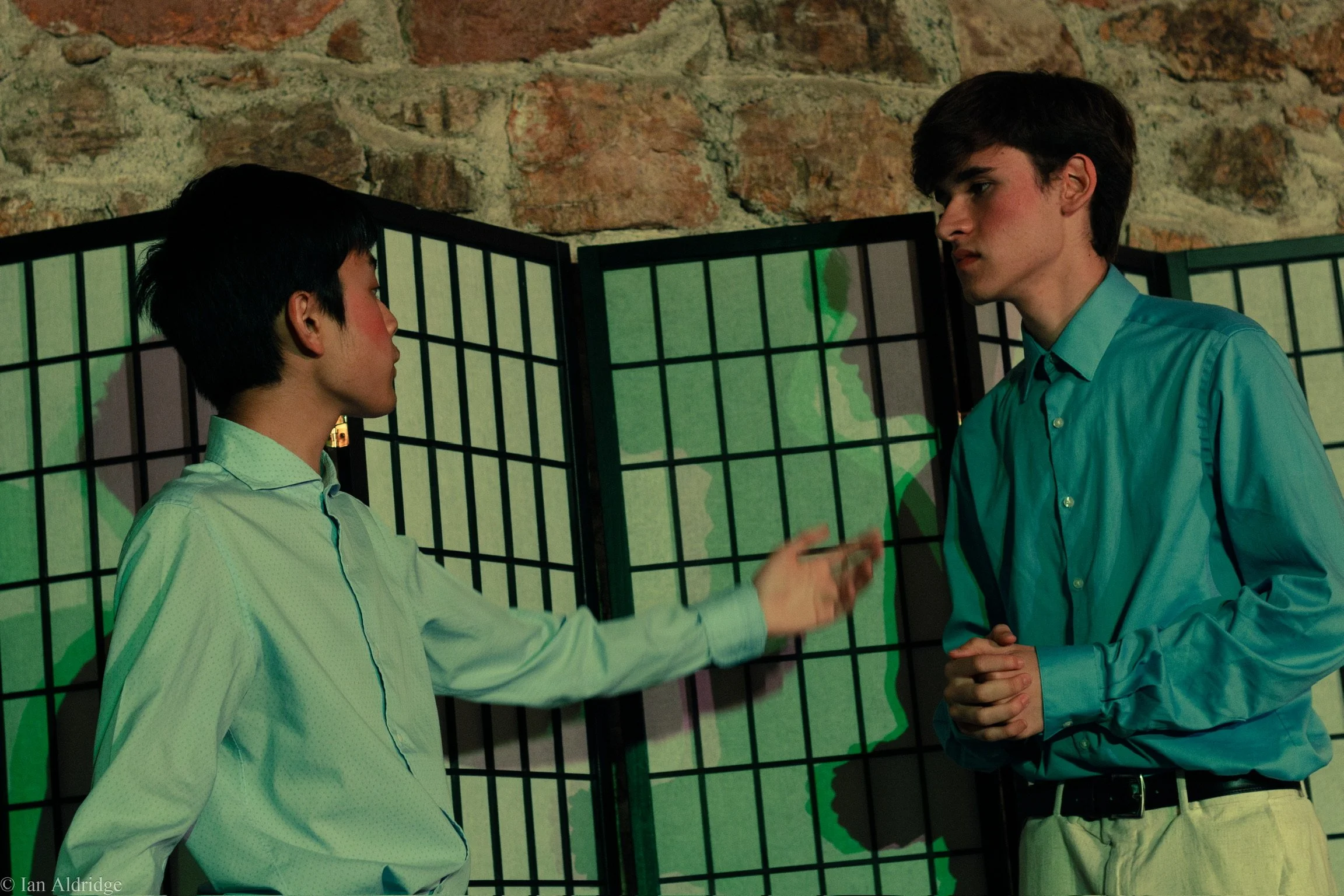 Two young men, one Asian and one Caucasian, standing close and engaging in conversation. They are dressed in collared shirts, with the Asian man gesturing with his hands. They are positioned in front of a decorative black folded screen with a stone w