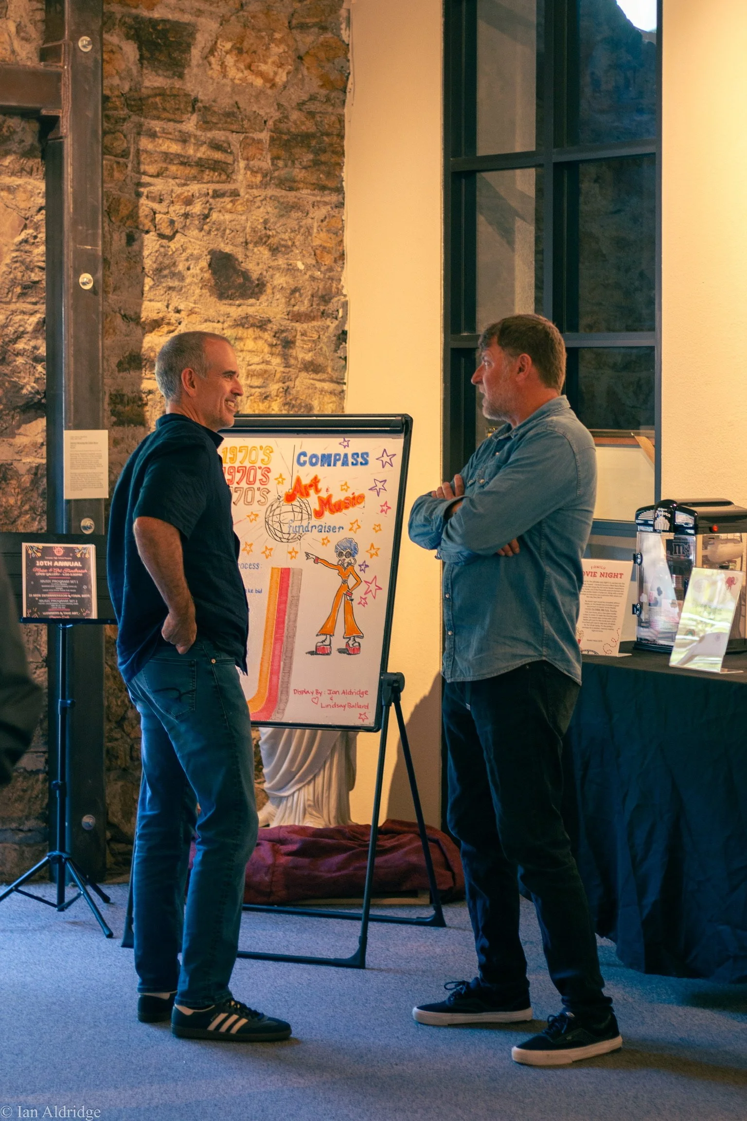 Two men standing and talking in front of a donation board at an indoor event with a stone wall background and a window.