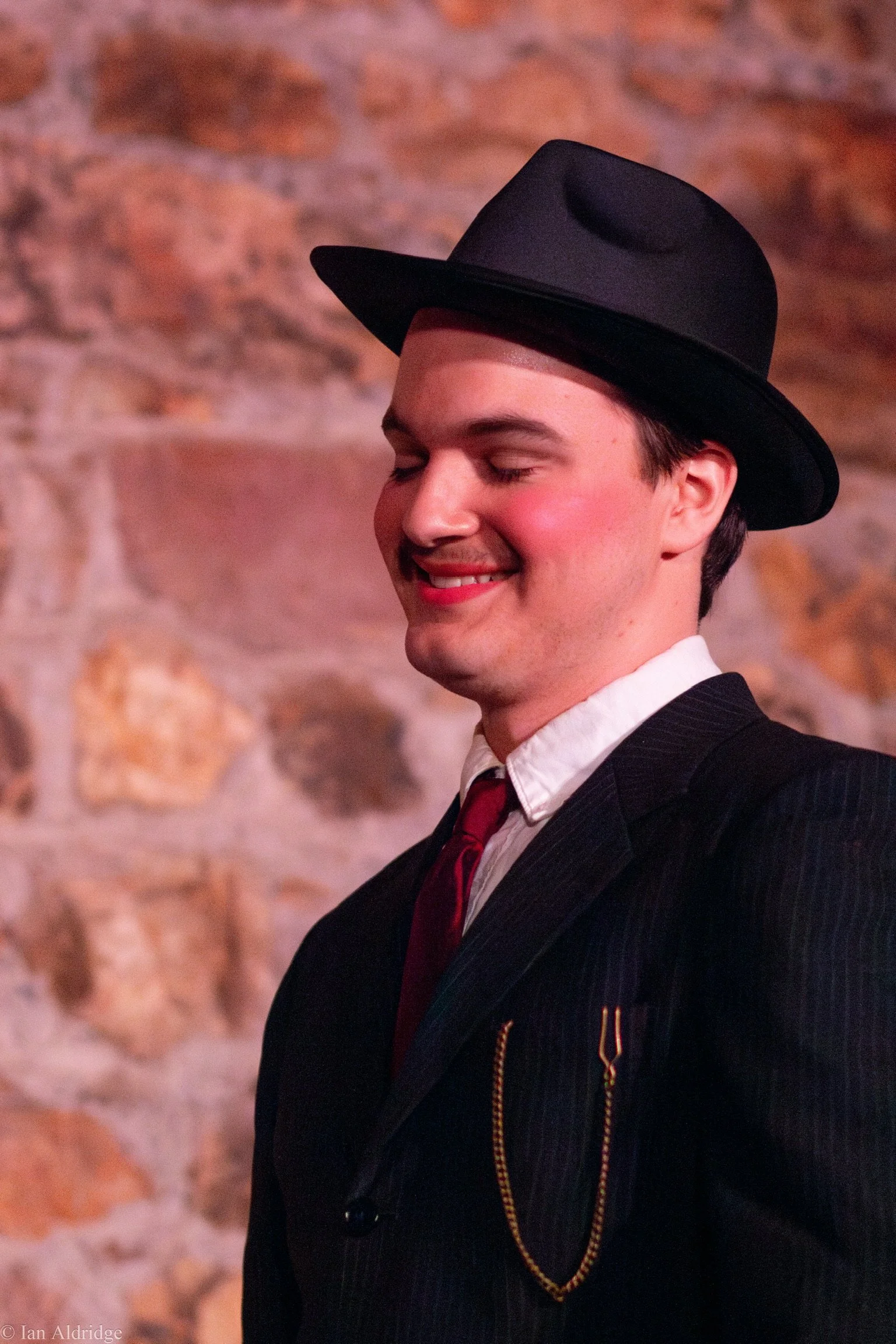 A man with closed eyes smiling, wearing a black hat, a dark suit, a white shirt, a red tie, and a gold chain, standing in front of a brick wall.