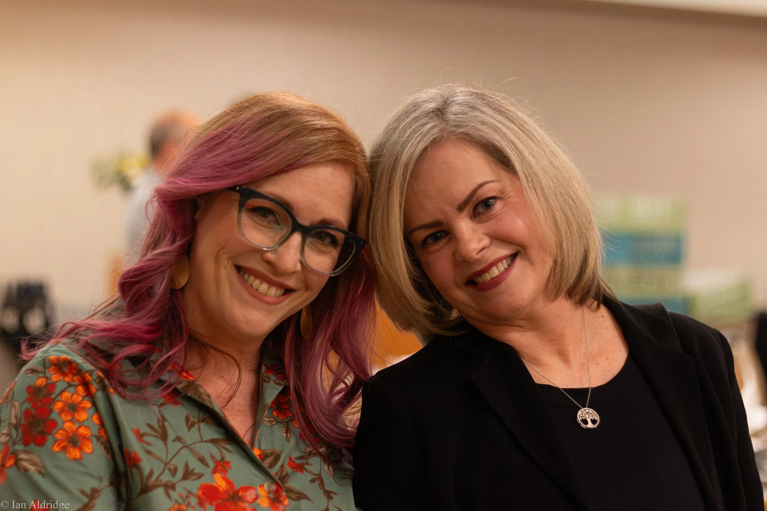 Two smiling women standing close together at an indoor event, with blurred background.