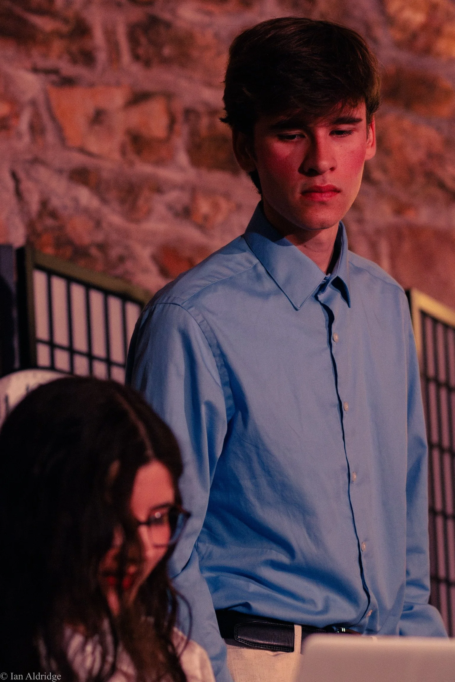 Young man with brown hair wearing a blue shirt, standing indoors against a brick wall, looking downward with a serious expression.