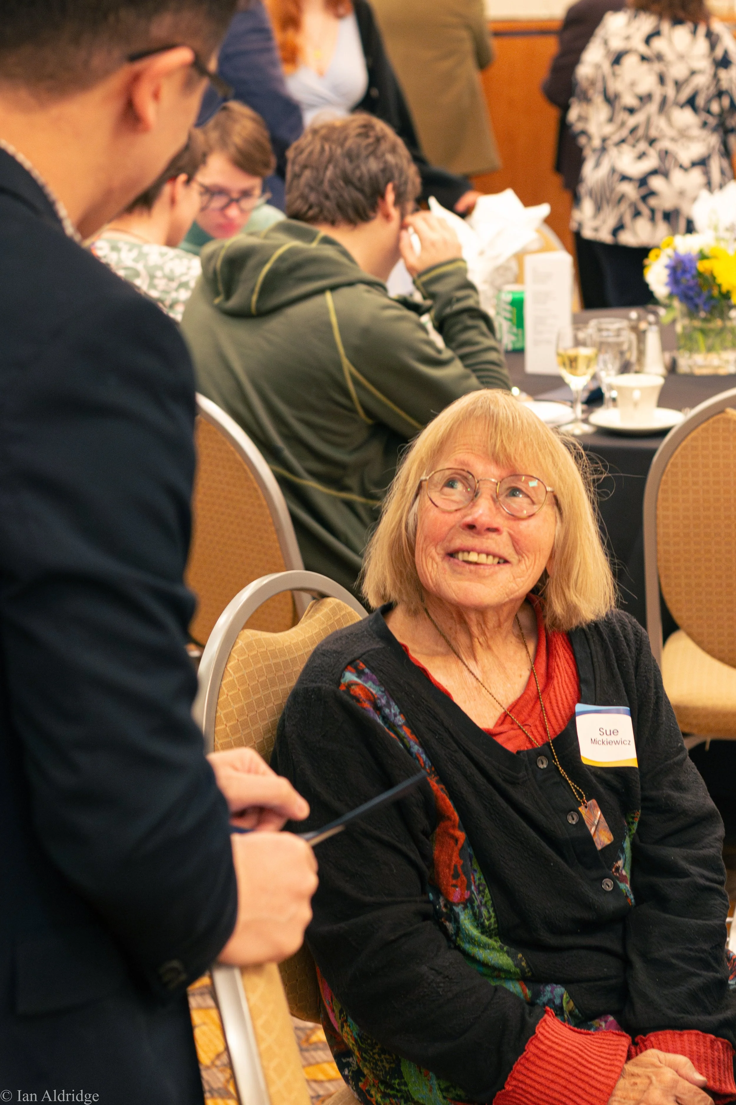 A woman with glasses and reddish hair, wearing a black cardigan with colorful patterns and a red shirt, is seated at a table with a name tag that reads 'Sue Mickiewicz.' She is smiling and looking up at a person standing nearby, who is holding a devi