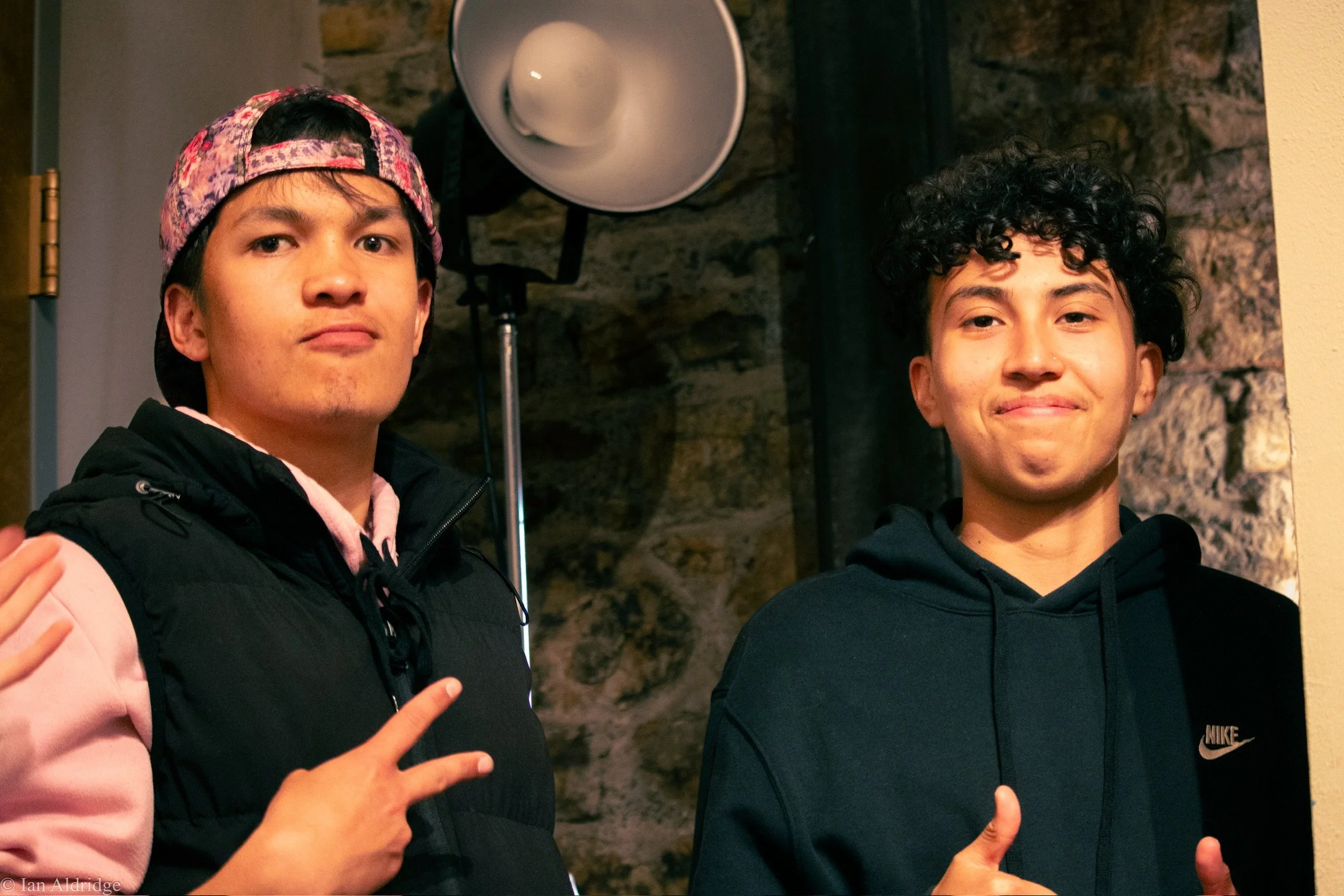 Two young men posing for a photo indoors, with one showing a peace sign and the other giving a thumbs-up. They are standing in front of a brick wall with a large lighting fixture visible.