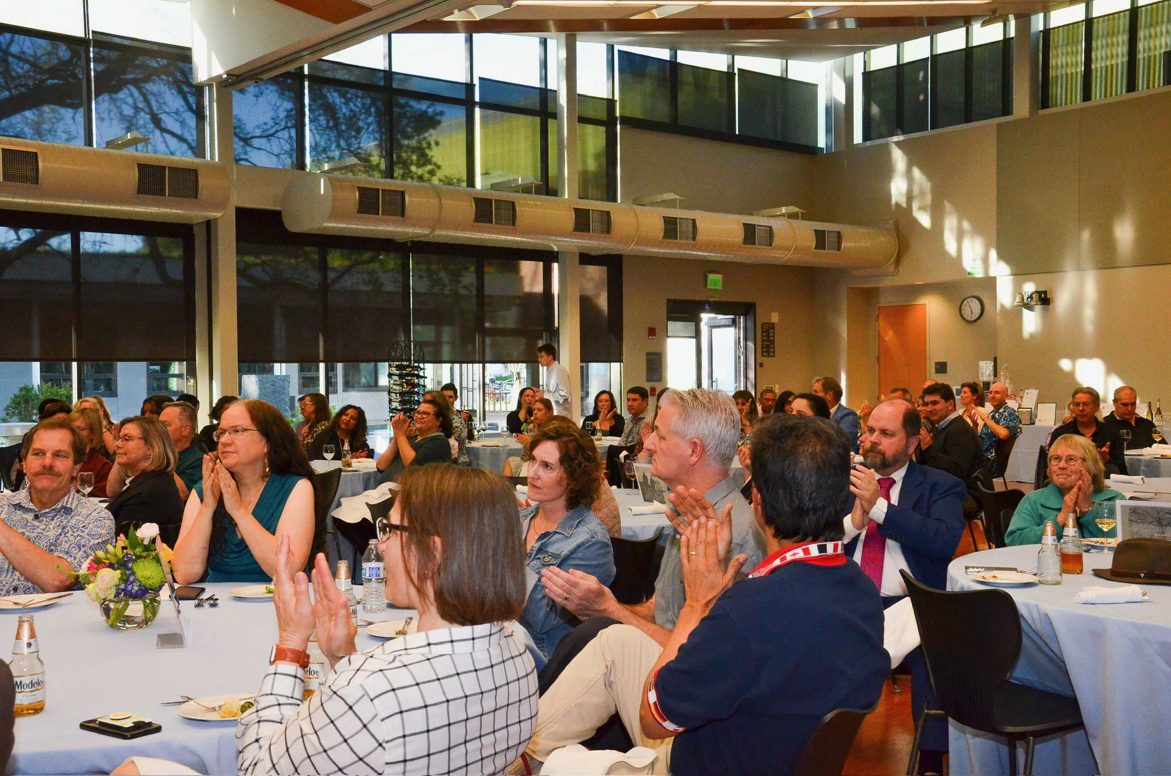 A large group of people seated at banquet tables, clapping and watching a presentation or speech in a spacious, modern room with large windows and high ceilings.