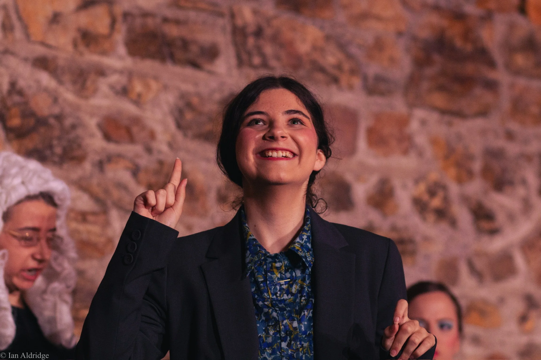 A young woman with dark hair, wearing a dark blazer and a floral shirt, smiling and gesturing with her index finger raised, in front of a brick wall. She appears happy and engaged in conversation.