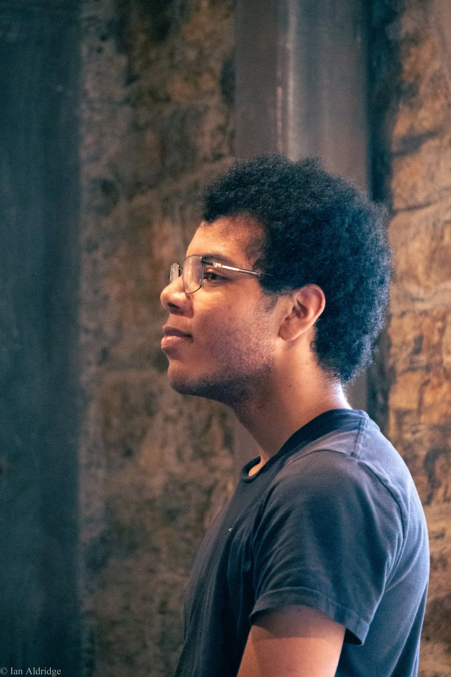 Profile of a young man with glasses, dark curly hair, wearing a dark T-shirt, standing against a textured brick wall.