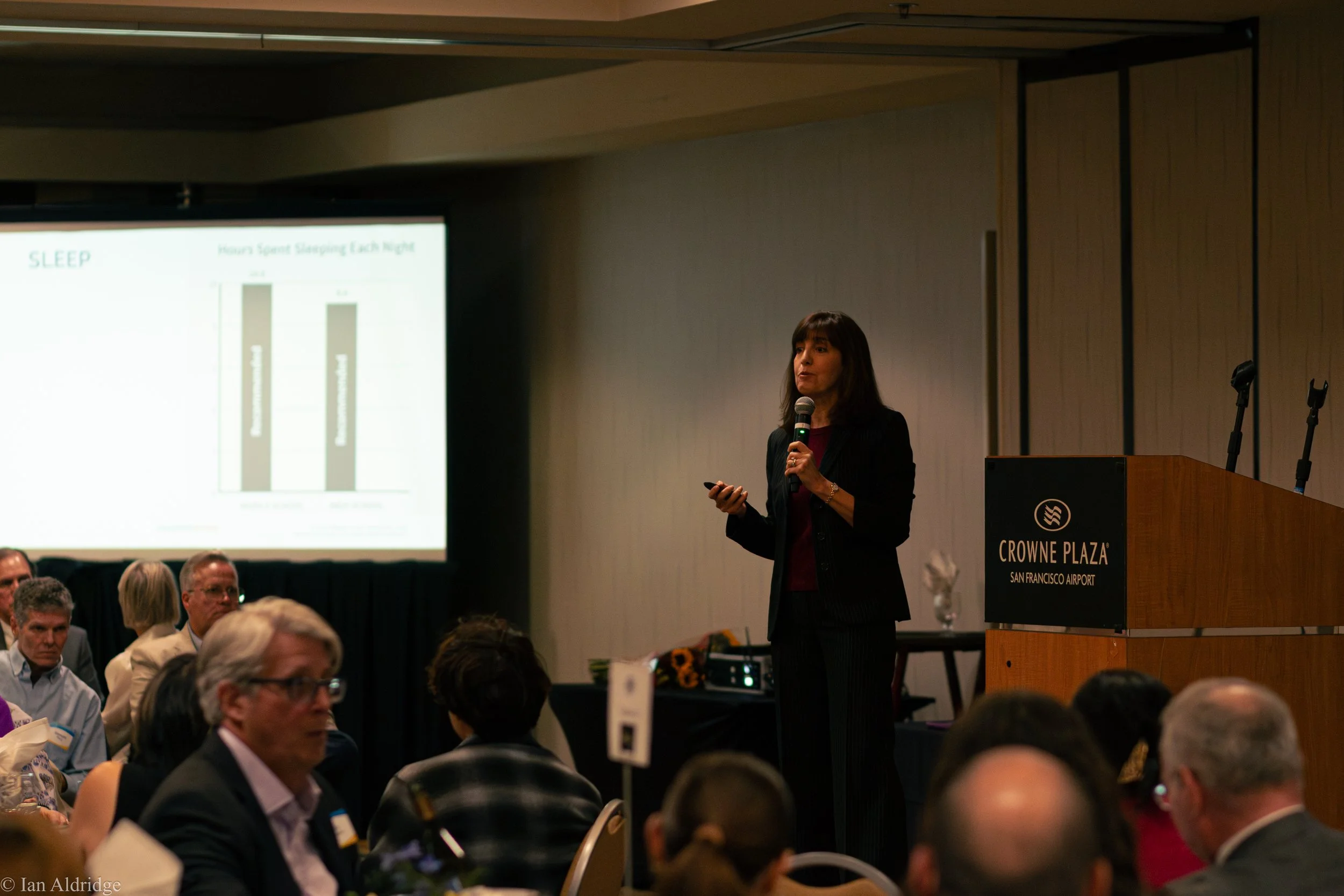 A woman giving a presentation at a conference in a ballroom with audience members seated and a screen displaying a slide about sleep on the left. The podium has a sign that reads 'Crowne Plaza San Francisco Airport'.