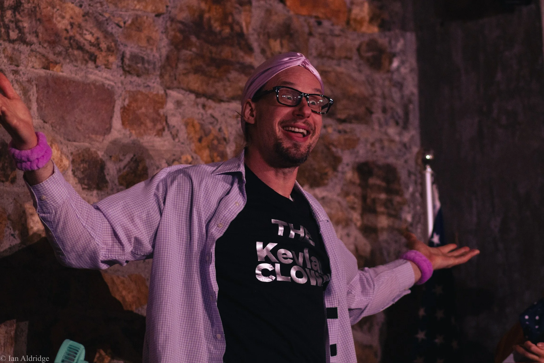 A man with glasses, a pink headscarf, and a checkered shirt smiling and raising his arms, standing against a brick wall in a dimly lit environment.
