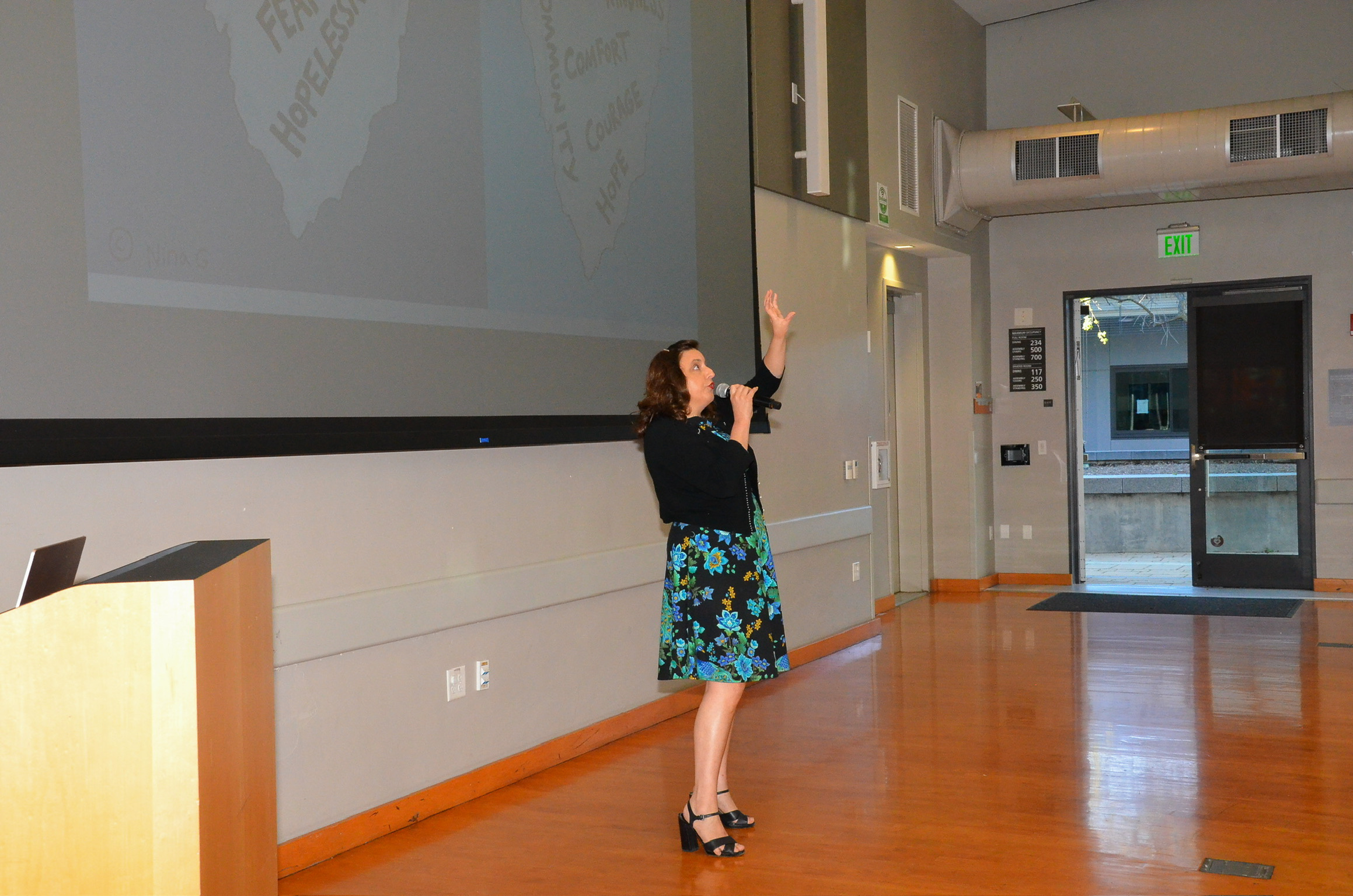 A woman wearing a black top and a blue floral skirt is giving a presentation in a room with wood floors. She is holding a microphone and gesturing upward with her left hand. Behind her, there is a large projection screen showing a map and text. The r