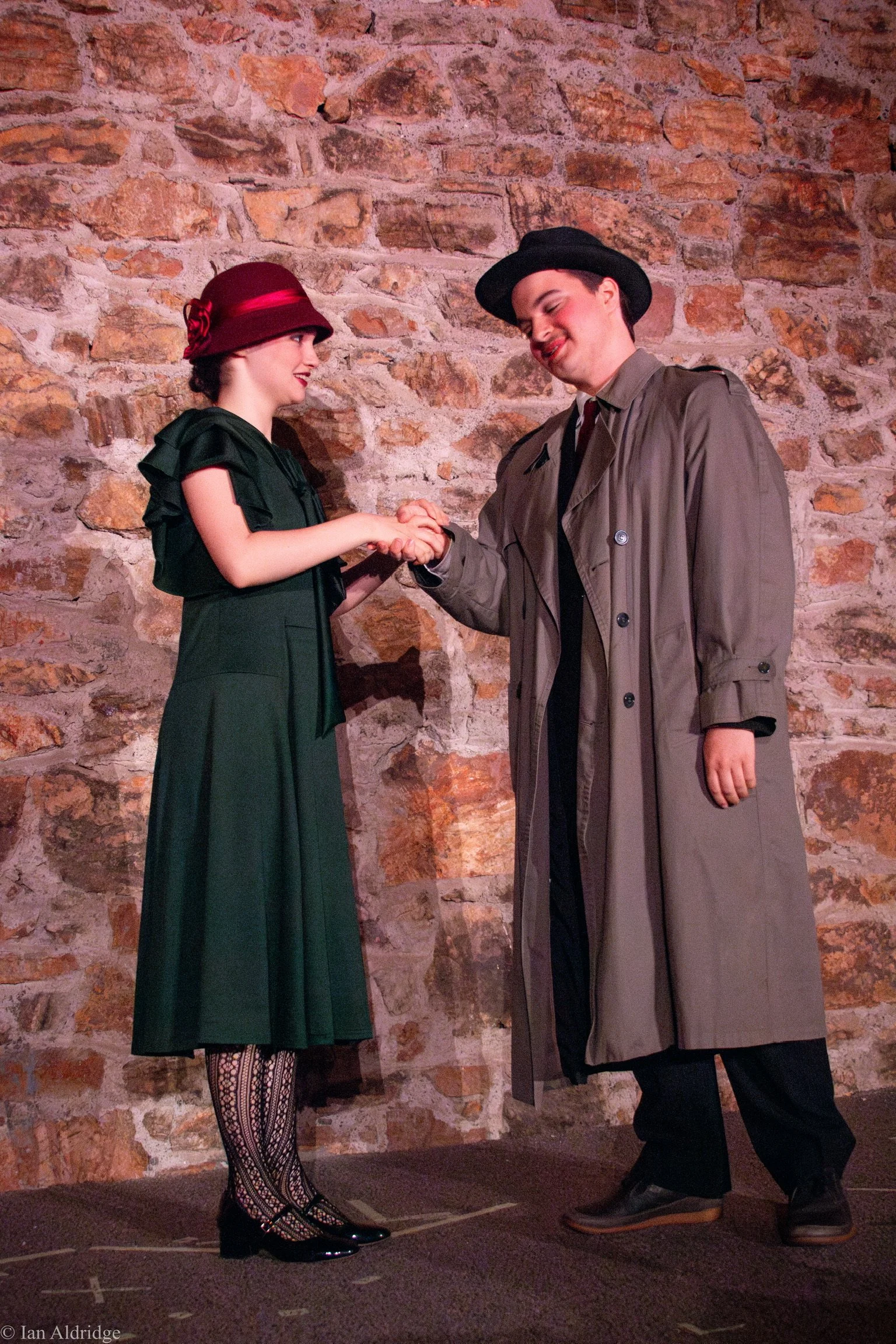 A man and woman shake hands on stage against a brick wall. The woman wears a dark green dress, decorative hat, and patterned tights. The man wears a trench coat and a wide-brimmed hat.