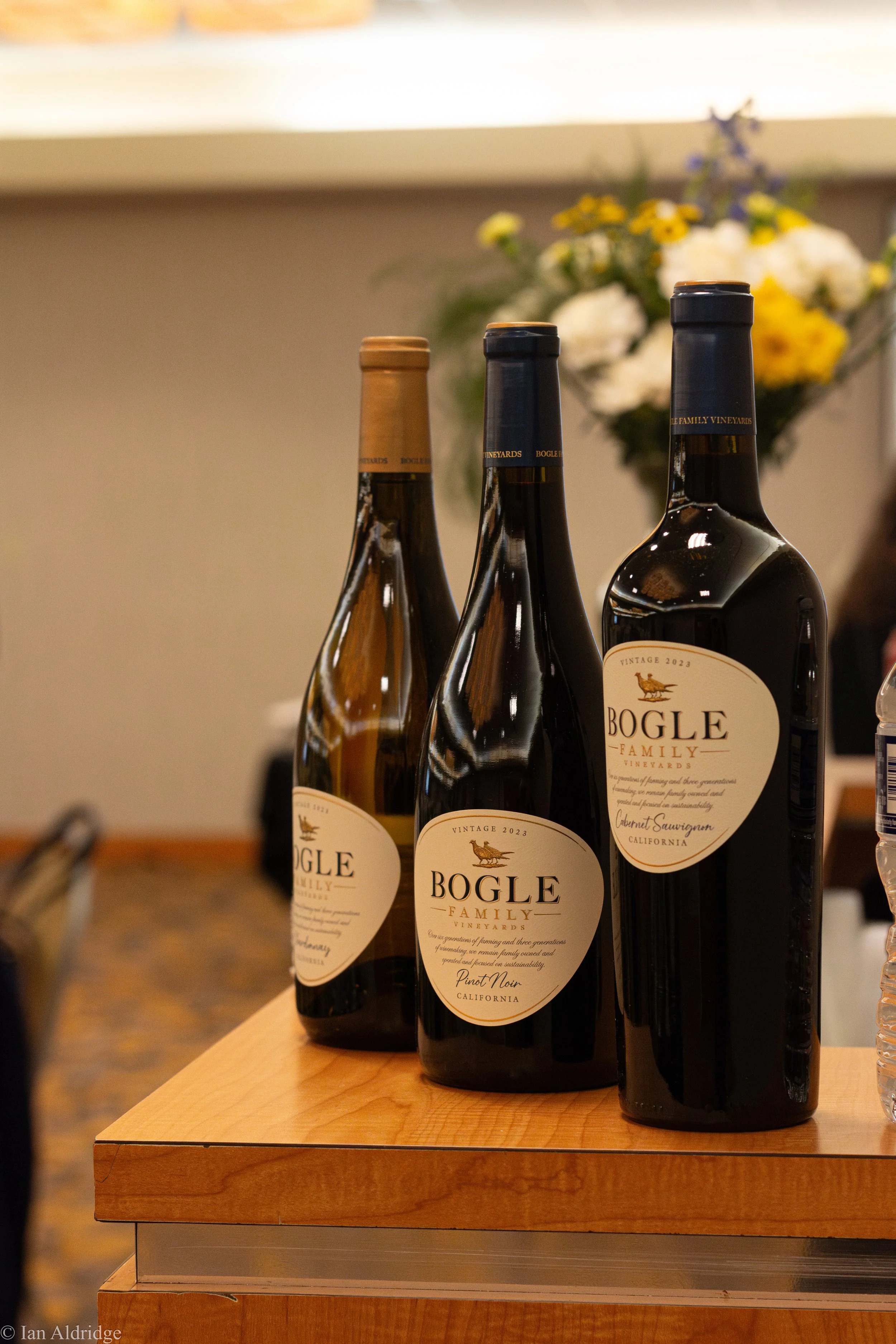 Three bottles of Bogle wine, including Pinot Noir, Cabernet Sauvignon, and others, are arranged on a wooden table with a floral arrangement in the background.