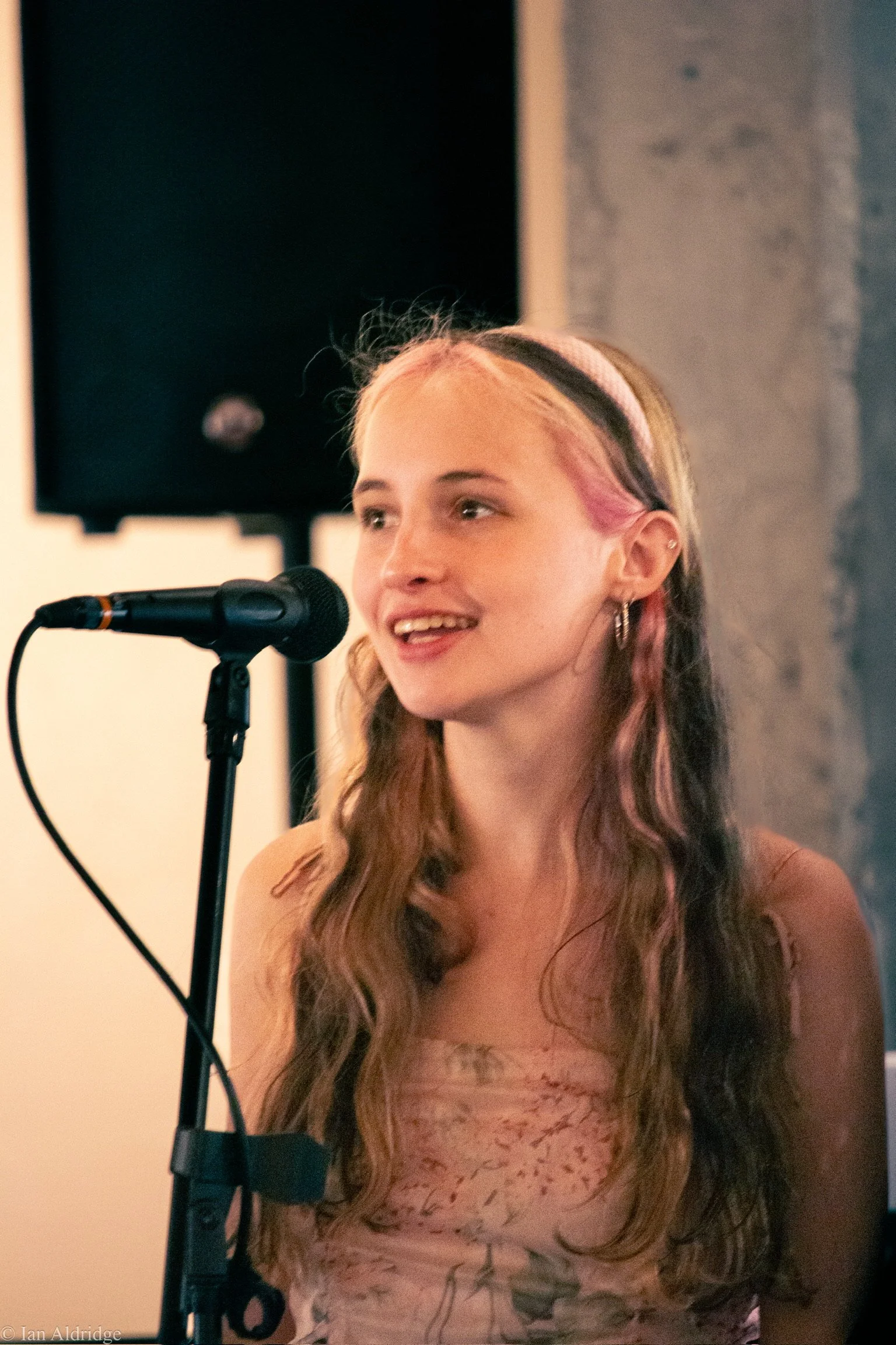 A young woman with long wavy hair, partially dyed pink, speaking into a microphone during a performance or speech. She is wearing a light-colored, patterned top and has multiple earrings.