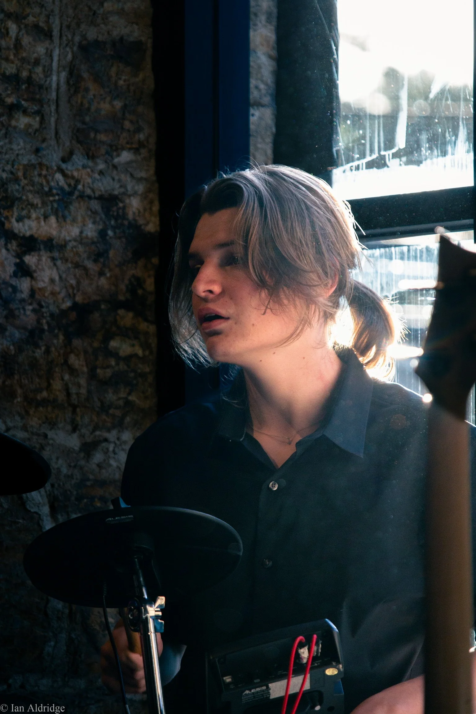 Young woman with shoulder-length hair working with electronic equipment near a stone wall and a window with bright sunlight outside.