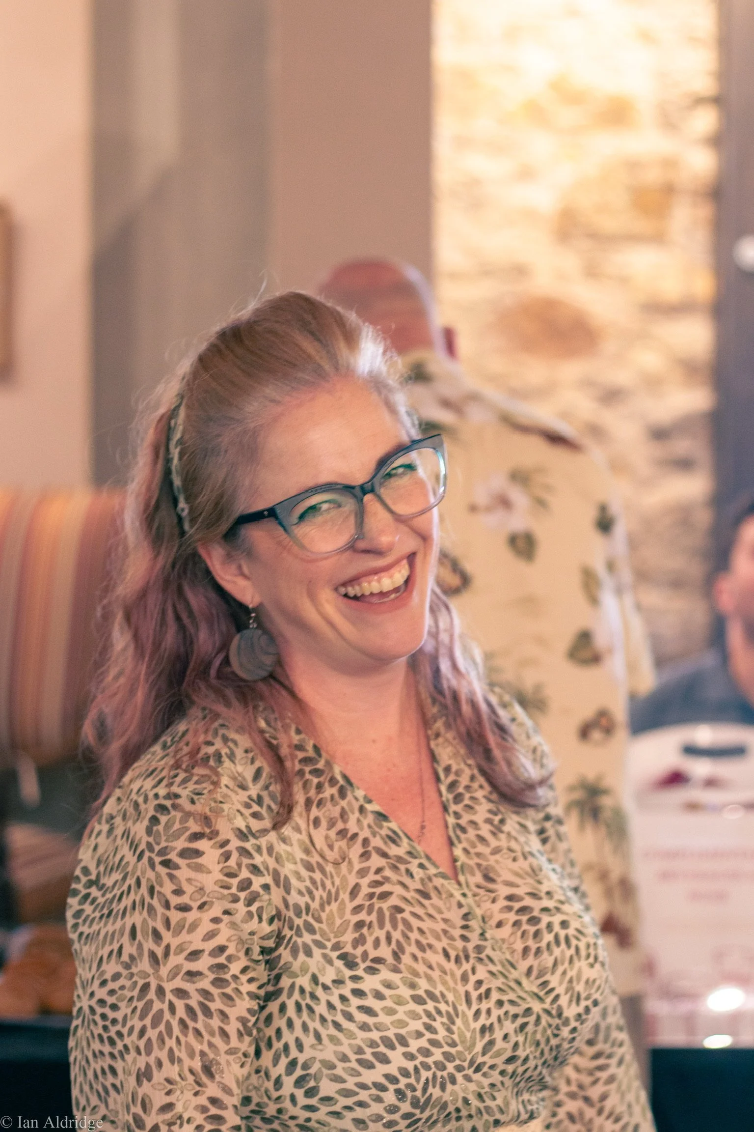 A woman with light skin, long wavy hair, wearing glasses and a leopard print blouse, smiling in an indoor setting.