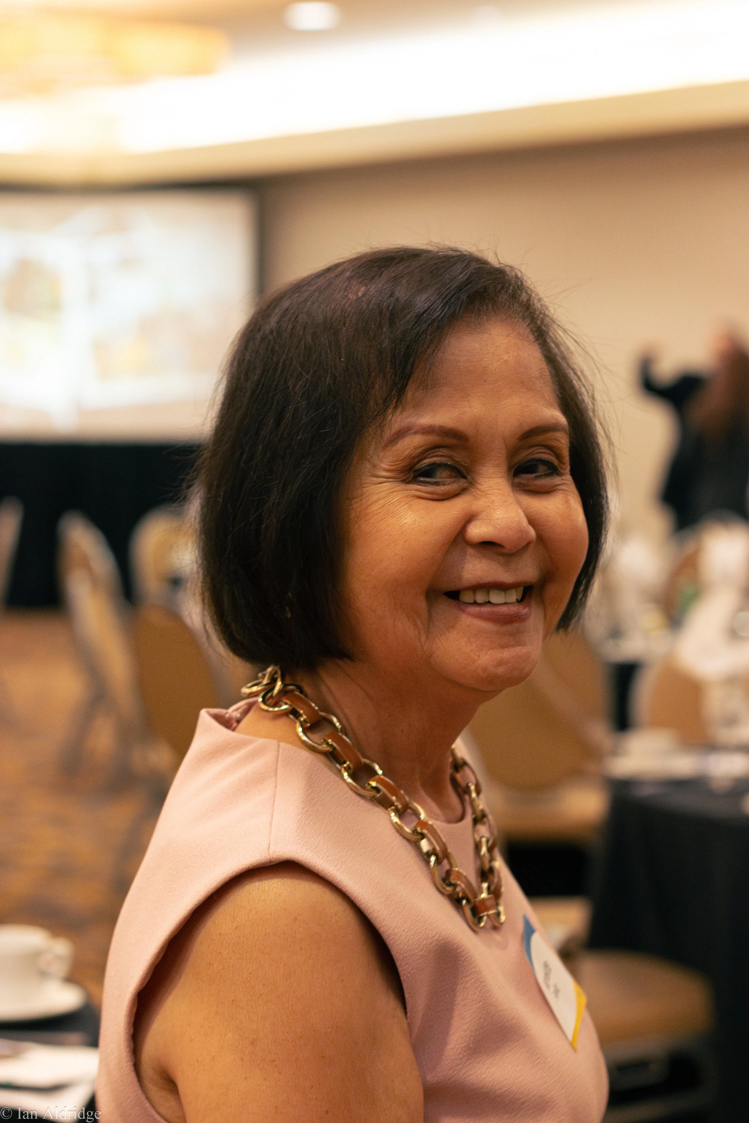 A smiling woman with short dark hair wearing a peach-colored dress and a large gold chain necklace at an indoor event, with tables and chairs and a blurred projection screen in the background.