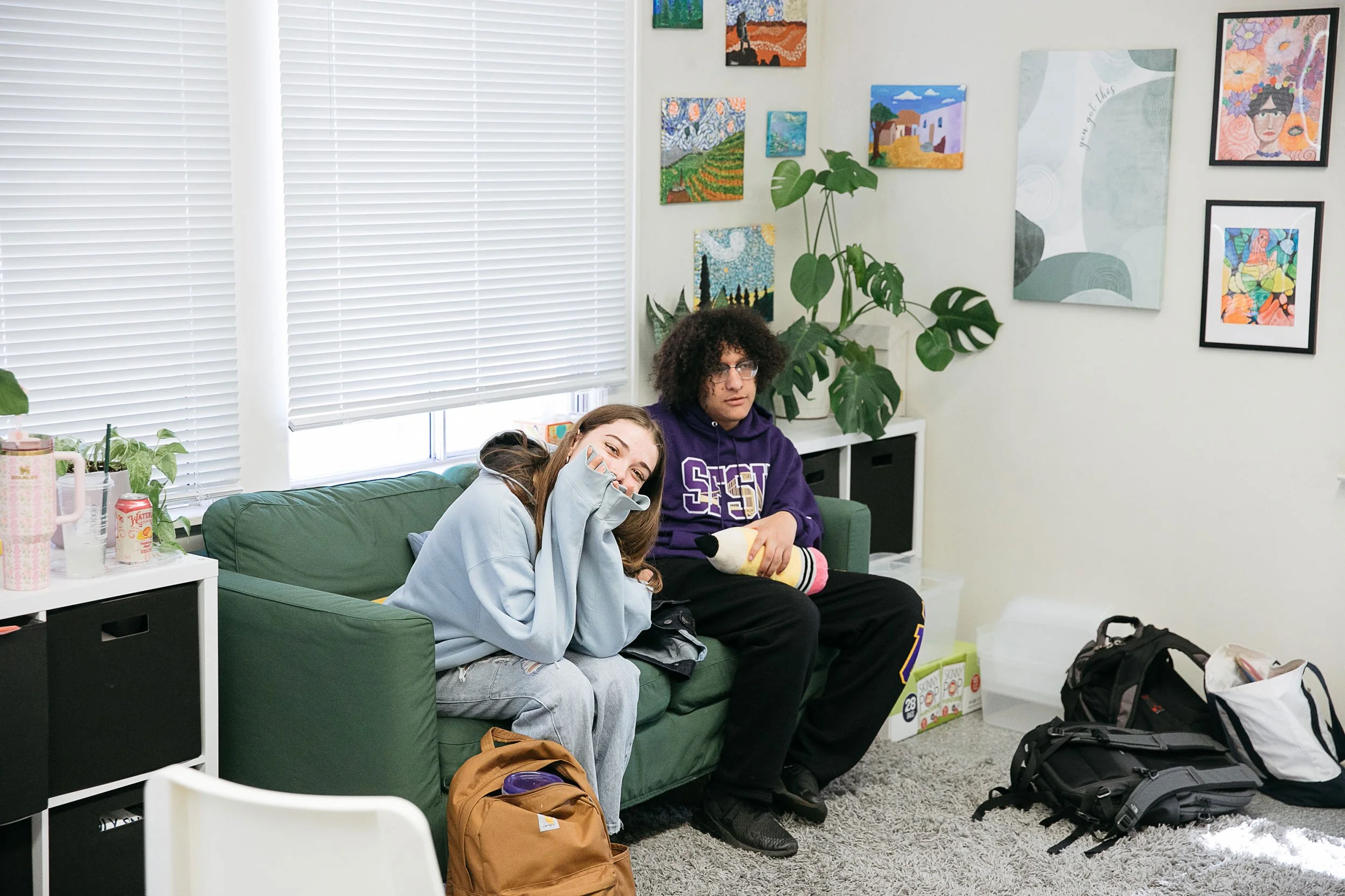 Two students sit on sofa in support office, one smiles at camera through fingers.