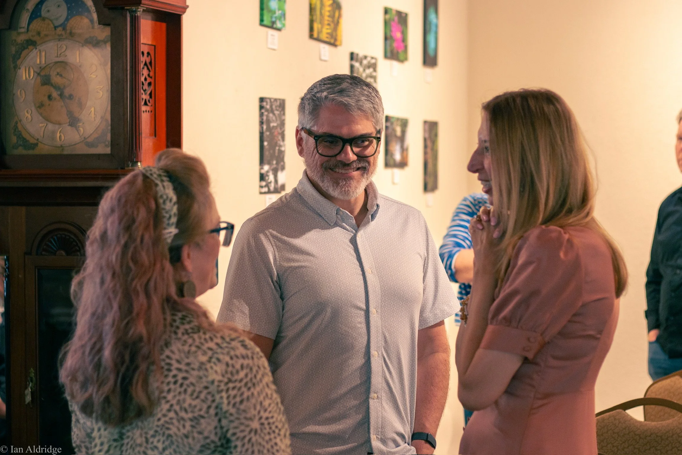 A group of people, including a man with glasses and gray hair, talking at an art gallery. The background features various colorful paintings and a vintage clock on a wooden cabinet.
