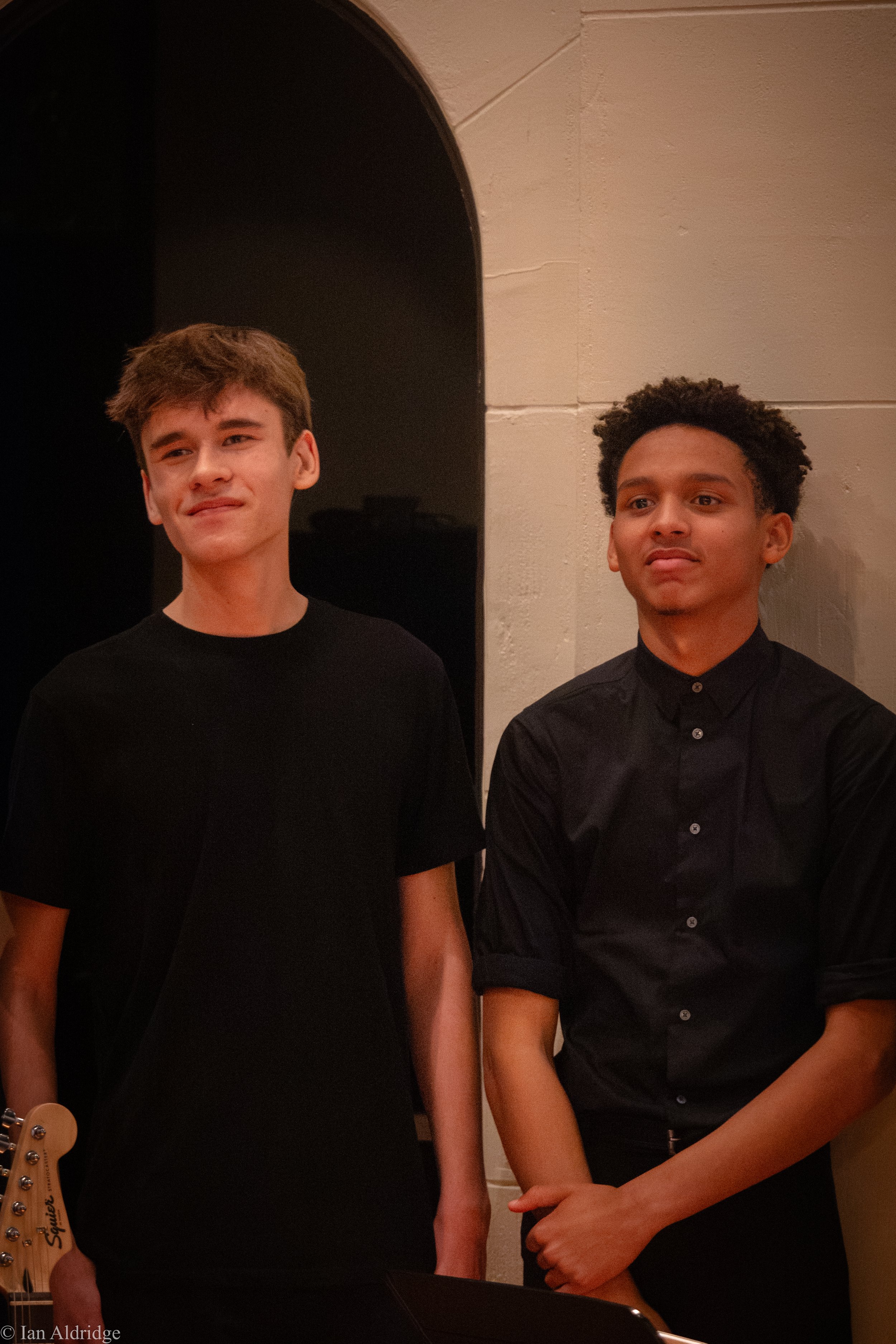 Two young men standing next to each other against a light-colored wall, both dressed in black, in a dimly lit room.