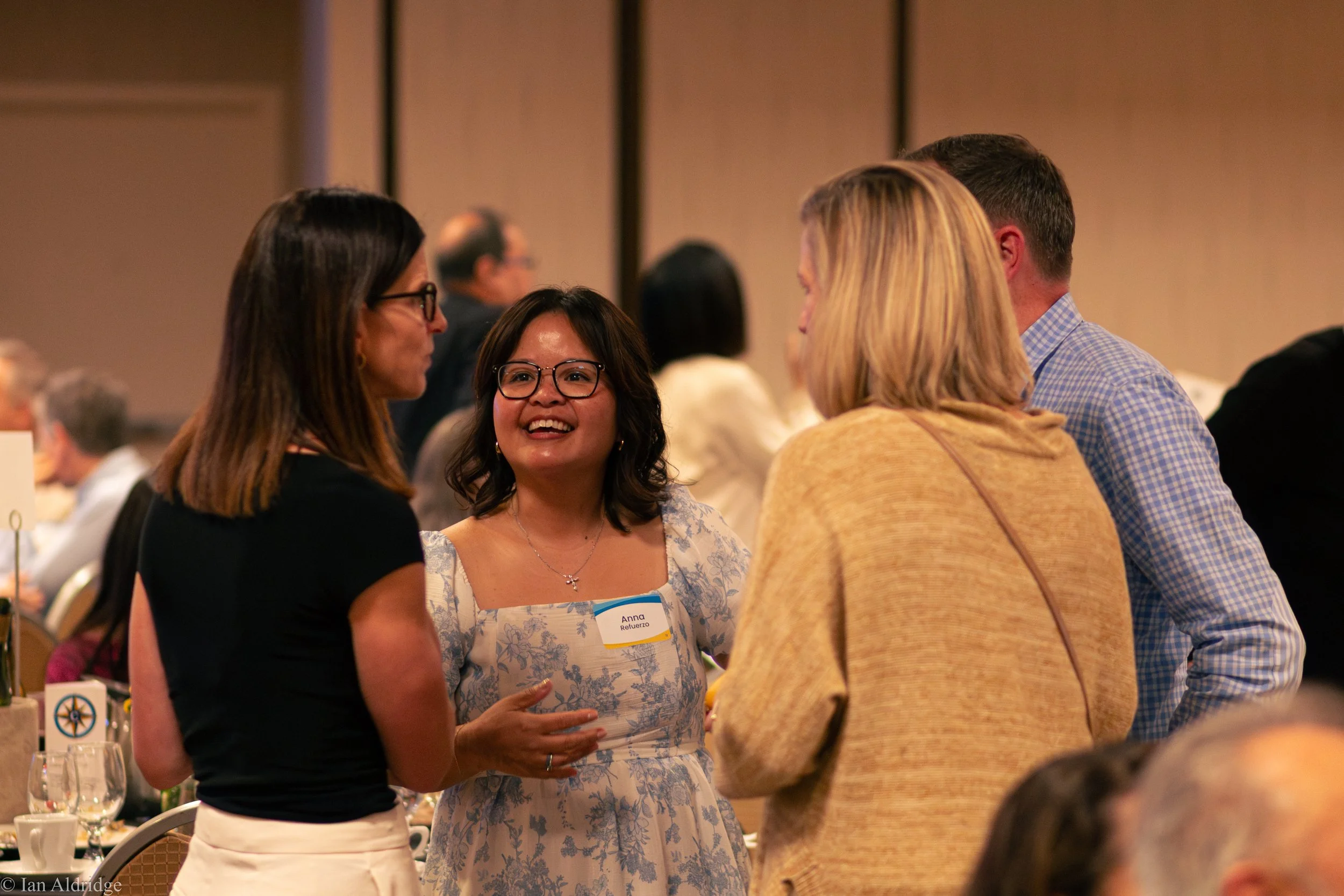 People engaged in conversation at a formal event or conference, with tables, glasses, and decorations visible in the background.