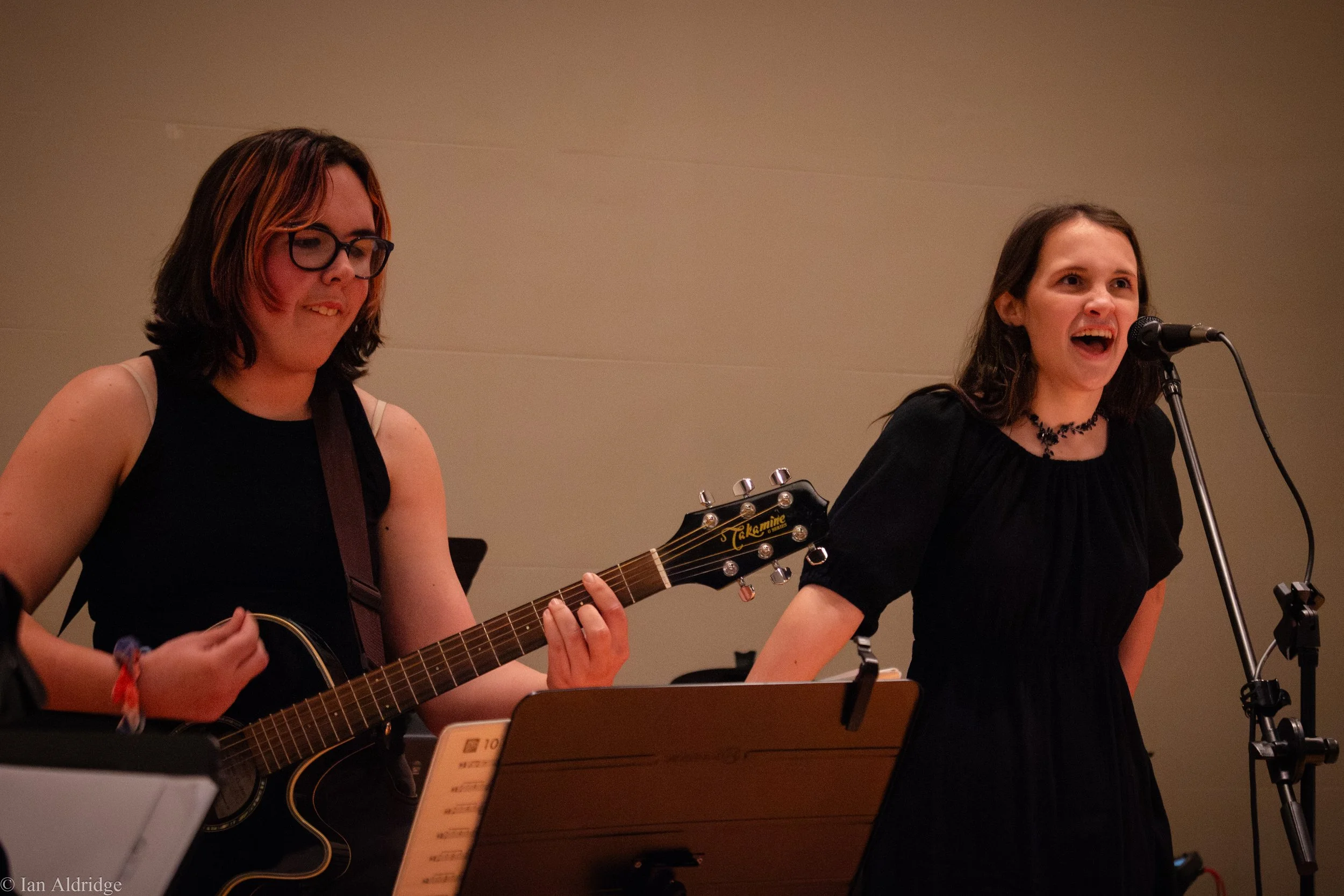 Two young women performing music with one singing into a microphone and the other playing an acoustic guitar on stage.