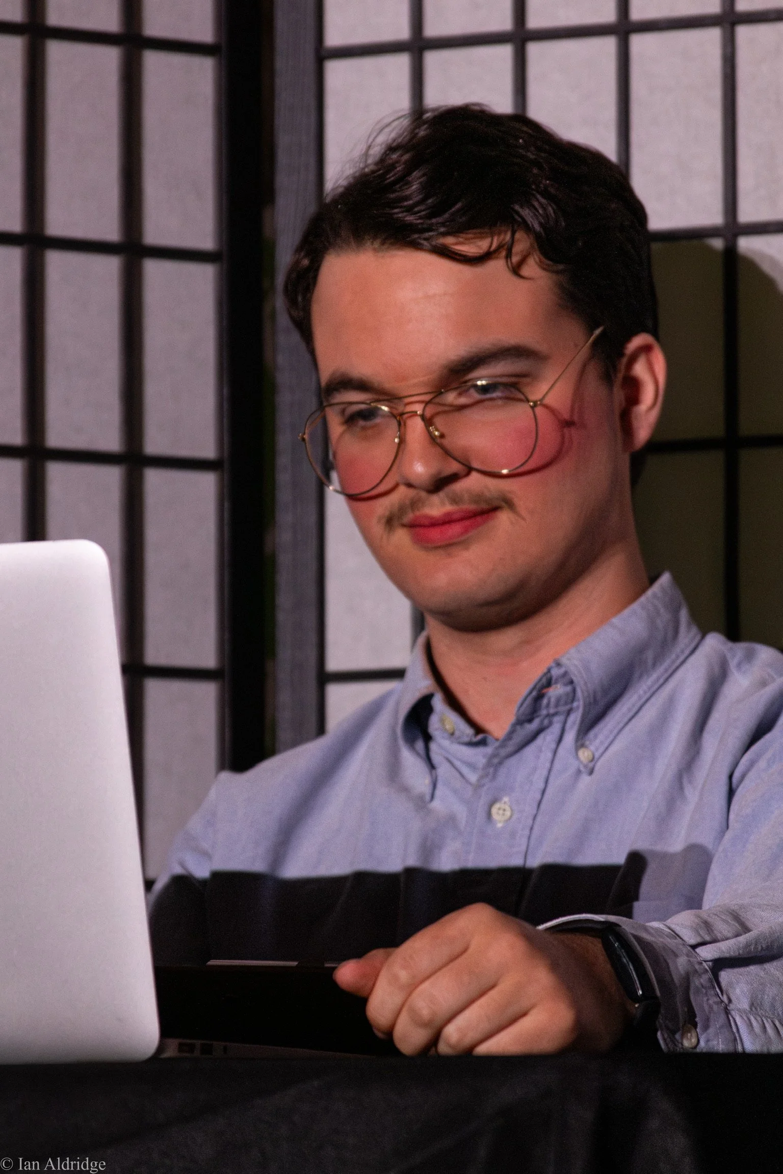 A young man with glasses and a mustache looking at a laptop screen, sitting at a table with a black tablecloth, in front of a decorative partition with a grid pattern.