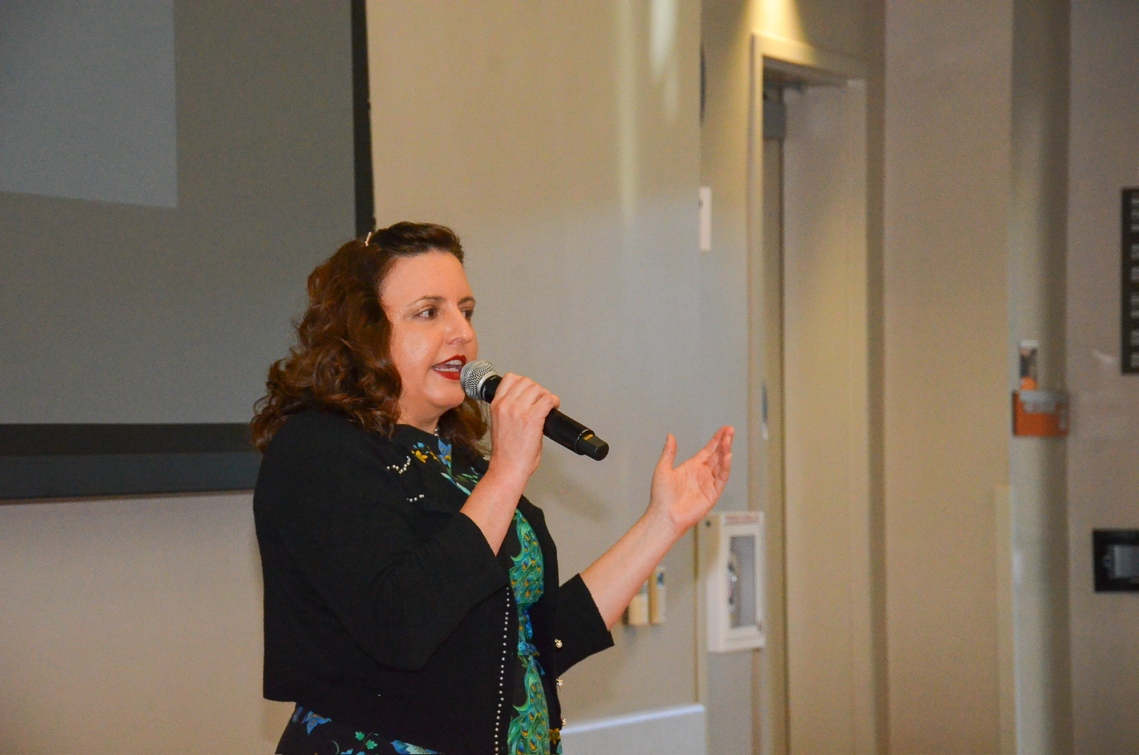 A woman speaking into a microphone during a presentation or speech in a room with a large screen and display panels.