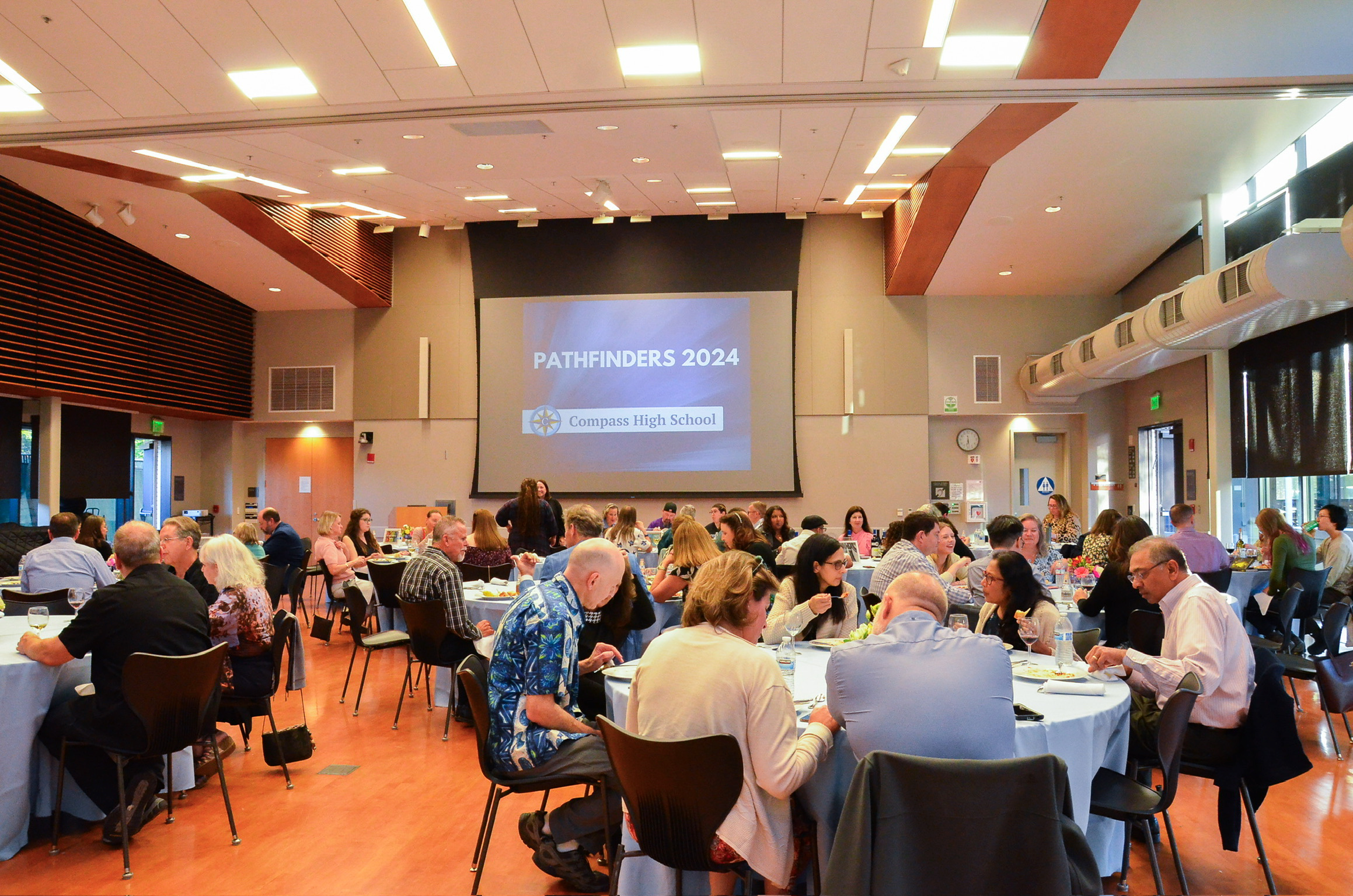 A large indoor room filled with round tables and many people attending a formal event or conference. There is a large screen displaying 'PATHFINDERS 2024' and 'Compass High School.' The room has high ceilings, modern lighting, and windows on one side