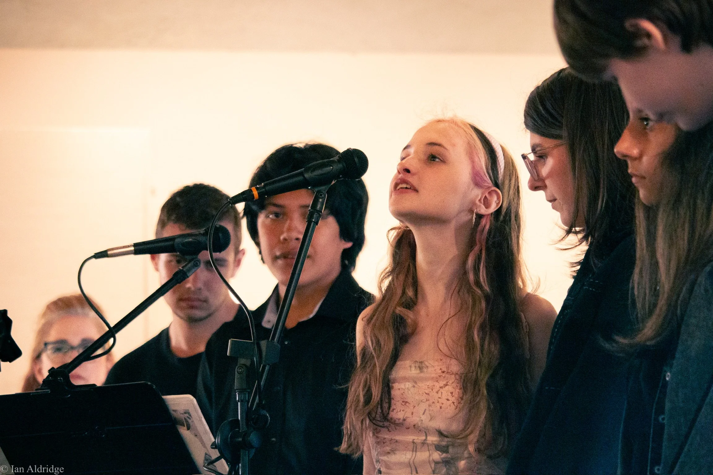 A group of young people standing together, some in front of microphones, likely at a performance or rehearsal. The girl in the center has long hair with pink tips and is looking slightly upwards. Others around her are focused or looking downward, pos