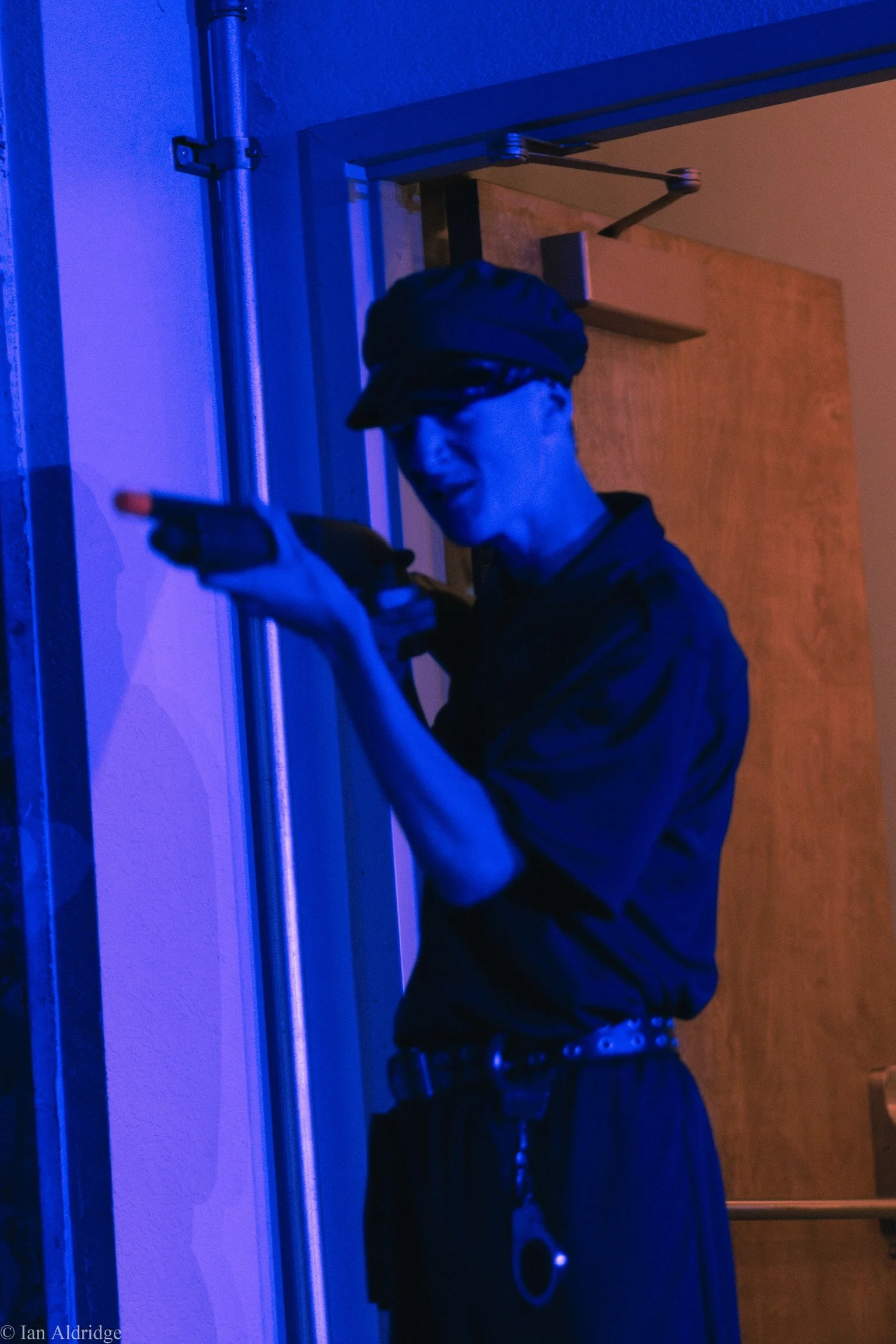 A person wearing a cap and dark clothing holding a gun in front of a blue-lit wall, with a wooden door in the background.