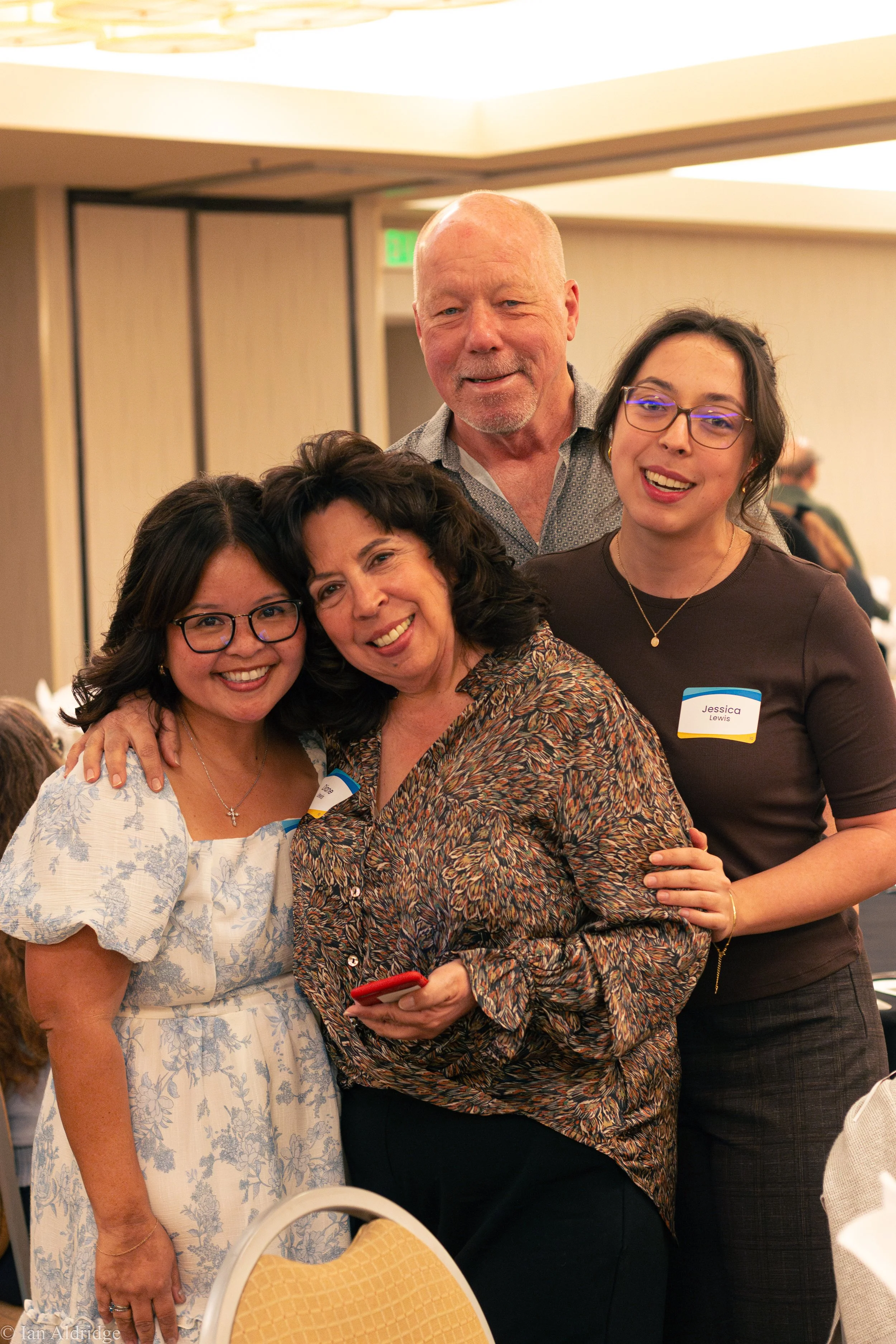 Group of four women at a social gathering, smiling and hugging, with name tags visible.