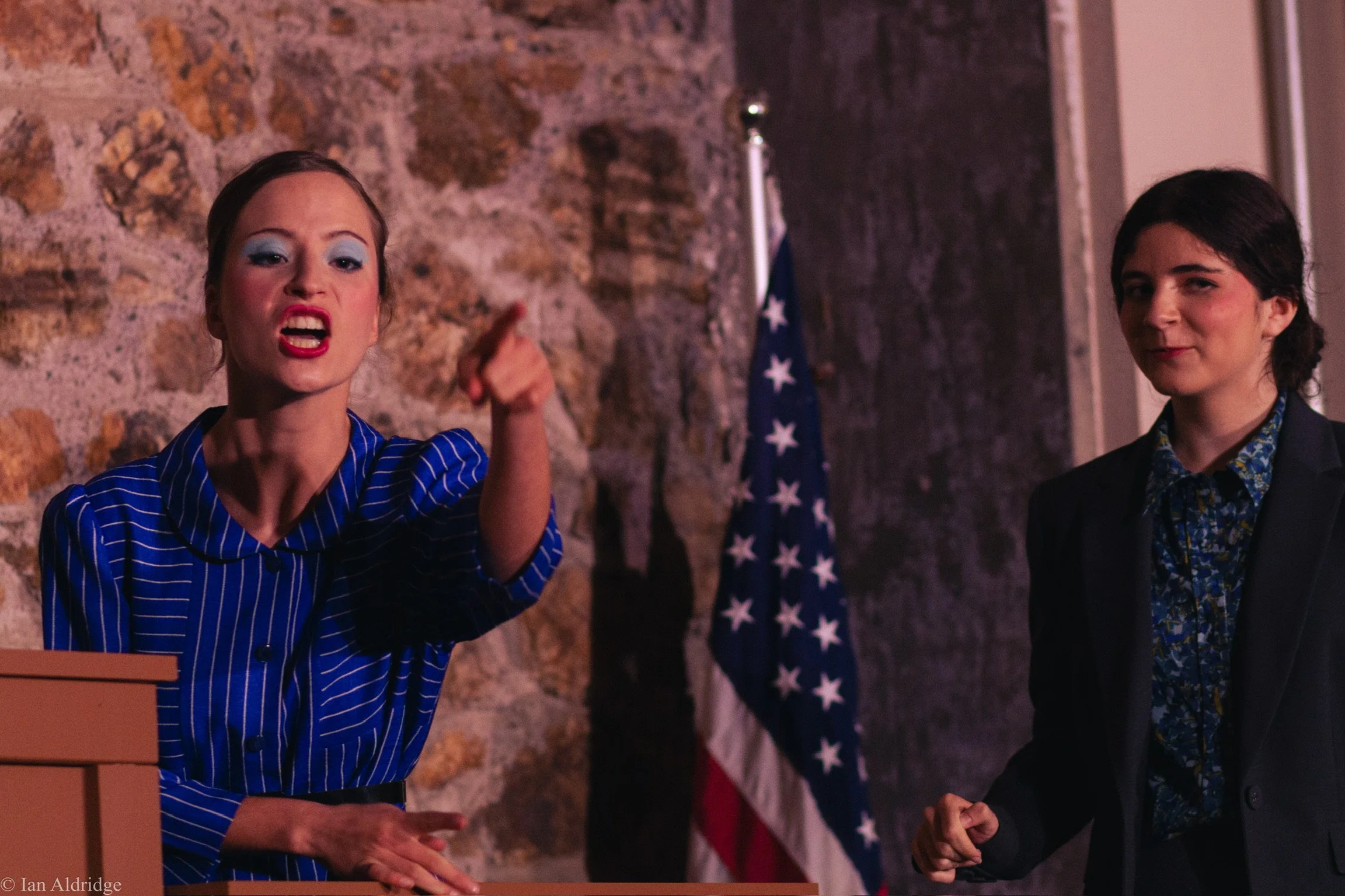 A woman with makeup and a blue pinstripe shirt is speaking passionately at a podium. Next to her stands a woman with dark hair, wearing a black blazer and a floral shirt, listening attentively. An American flag is visible in the background along with