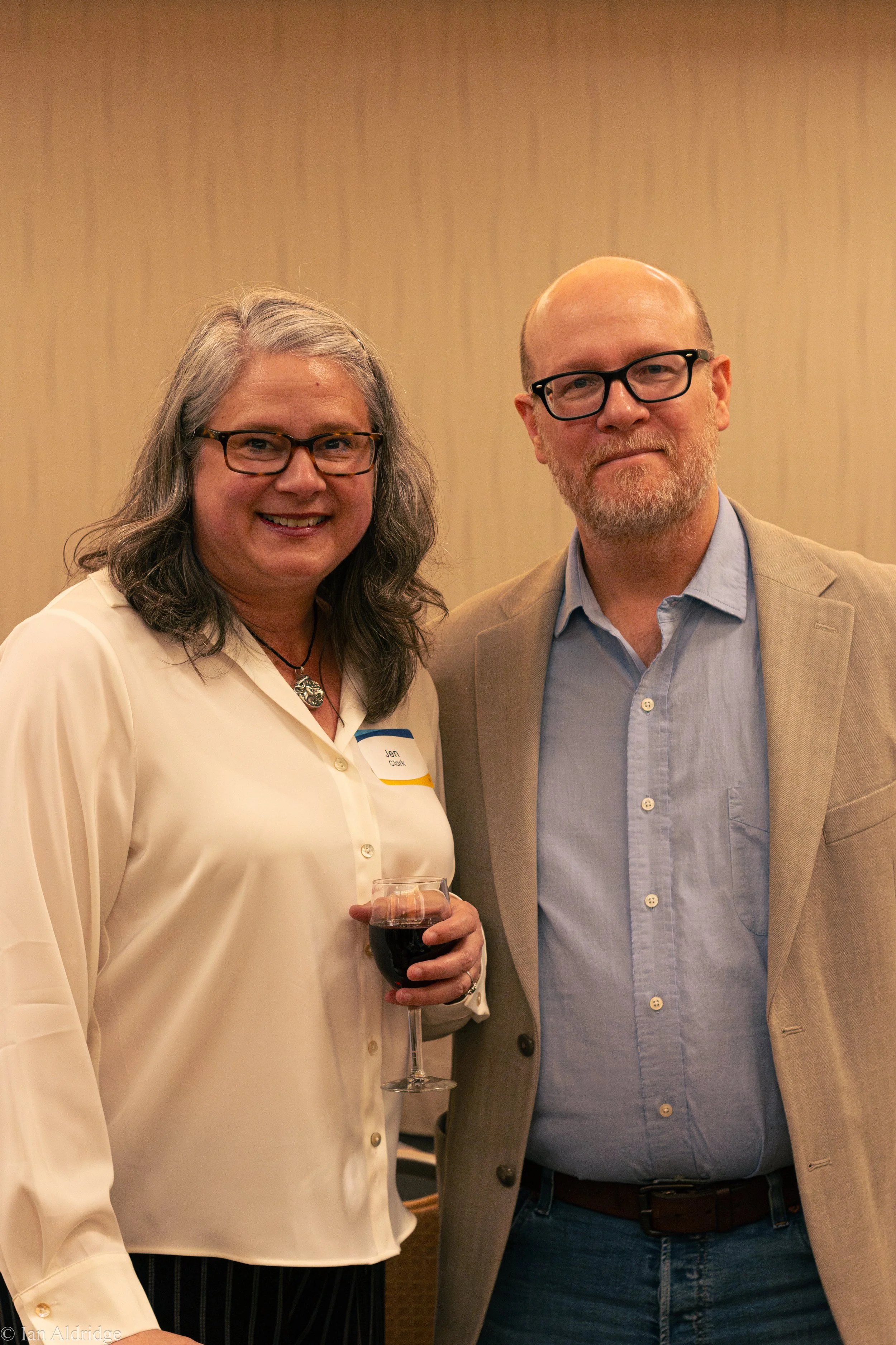 A woman and a man standing together at an event, smiling. The woman is holding a glass of red wine, and the woman has gray hair, glasses, and a cream-colored blouse. The man is wearing glasses, a beige blazer, a light blue shirt, and jeans.