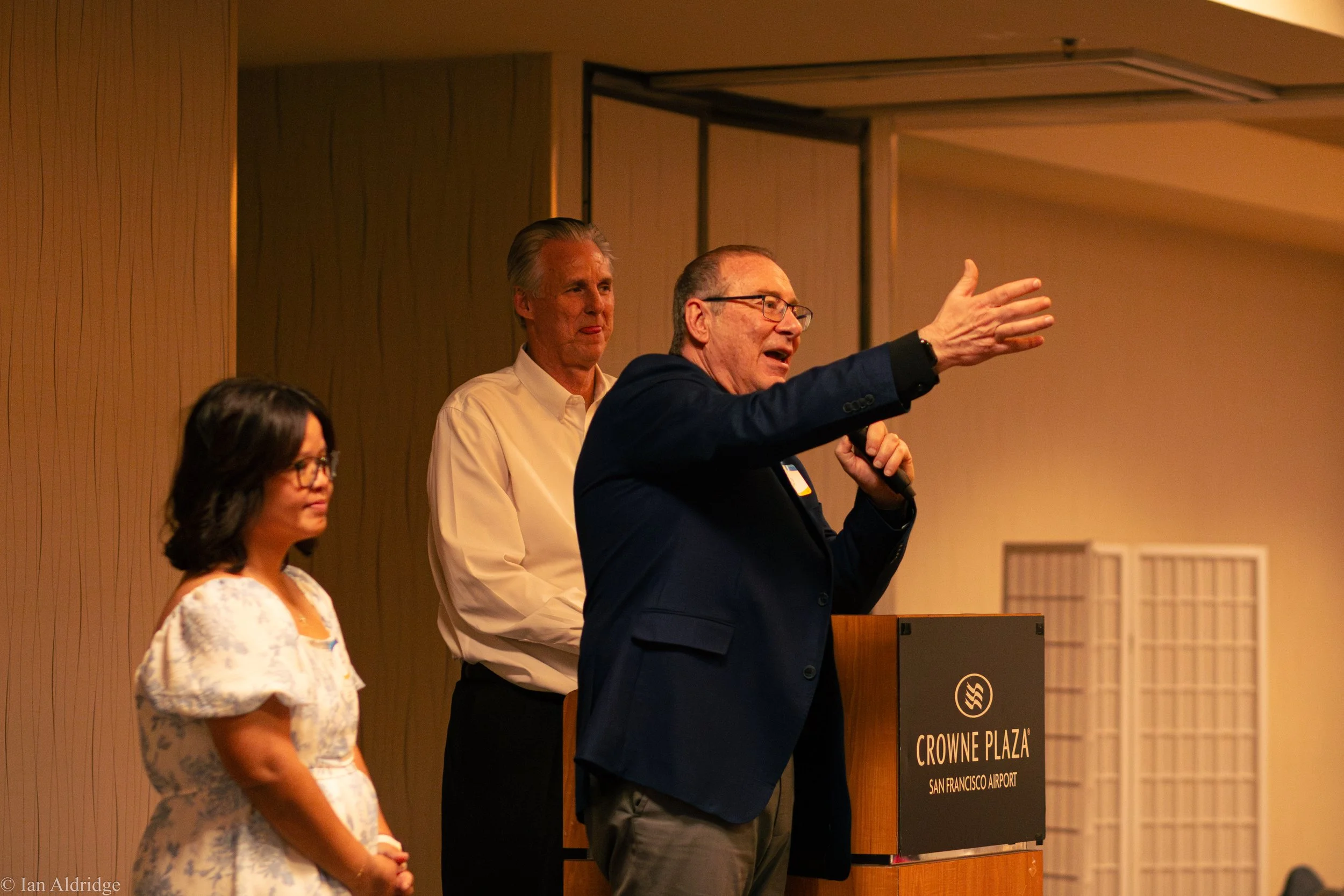 A speaker at a podium giving a presentation at Crowne Plaza San Francisco Airport, with two other women standing nearby, in a conference room with wood-paneled walls.