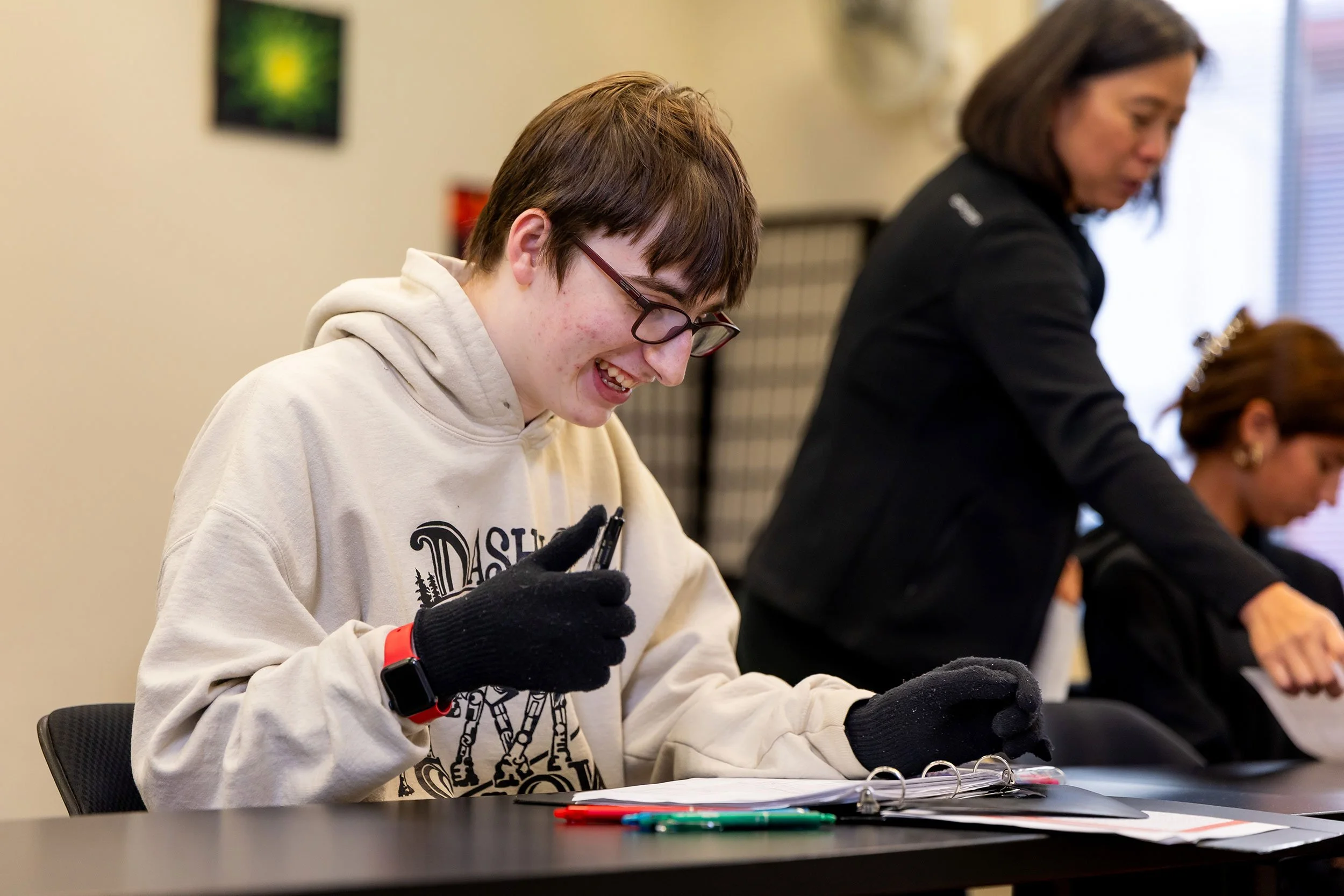 Smiling student works in classroom while wearing sensory gloves, and gives thumbs up.
