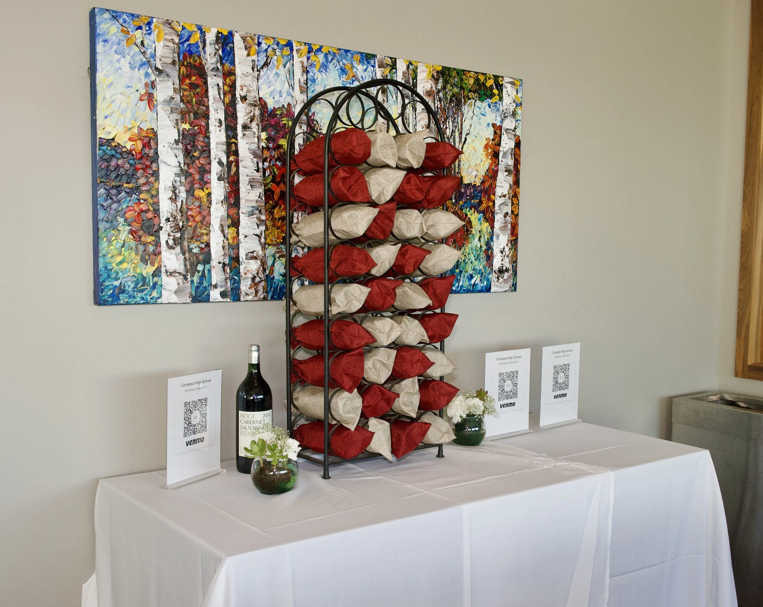 A display table featuring a wine bottle, decorative vases with flowers, and QR code cards in front of a vibrant, abstract forest painting on a wall.